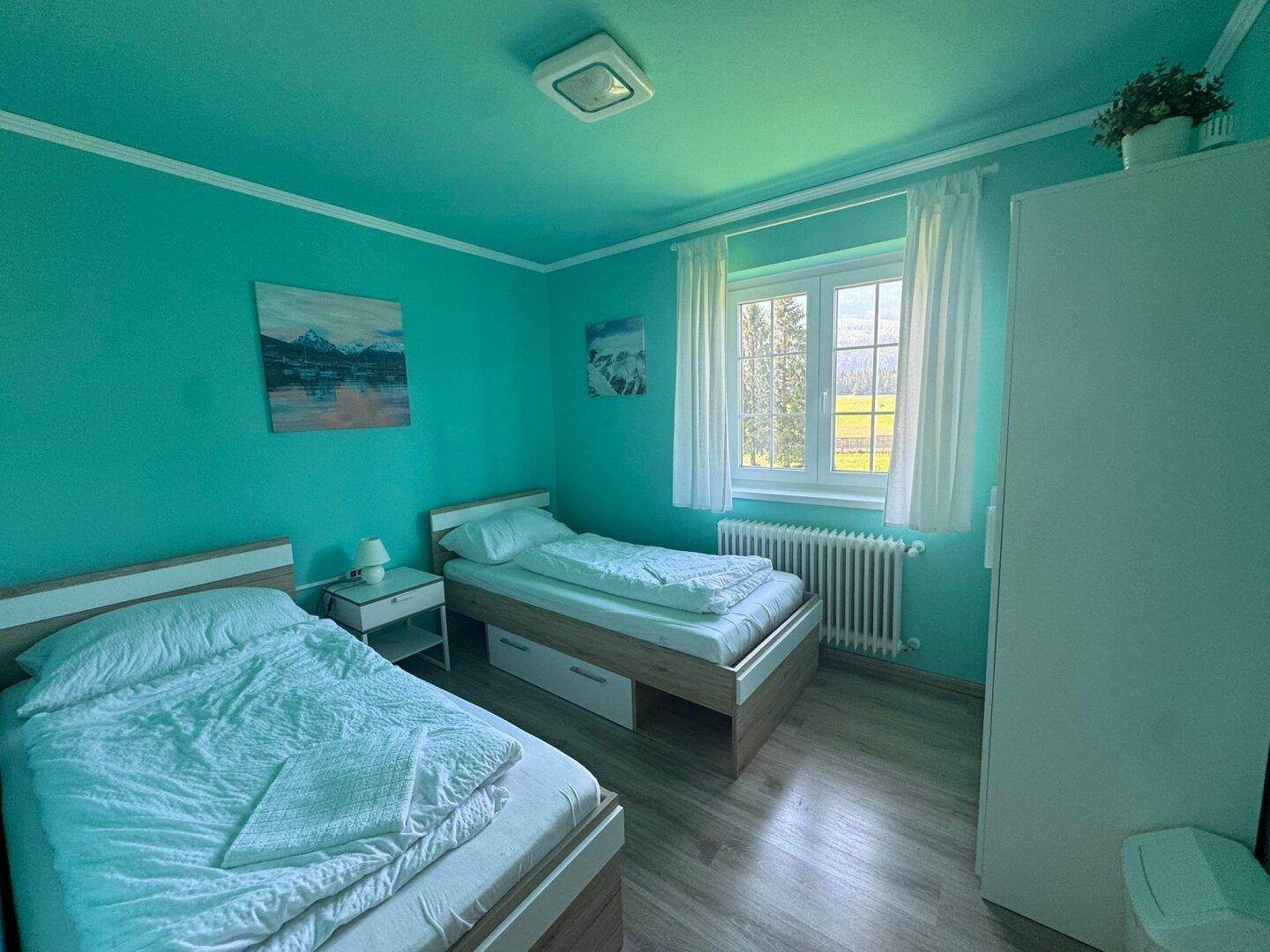 Freundliches Schlafzimmer mit zwei Einzelbetten, türkisfarbenen Wänden und Blick ins Grüne.