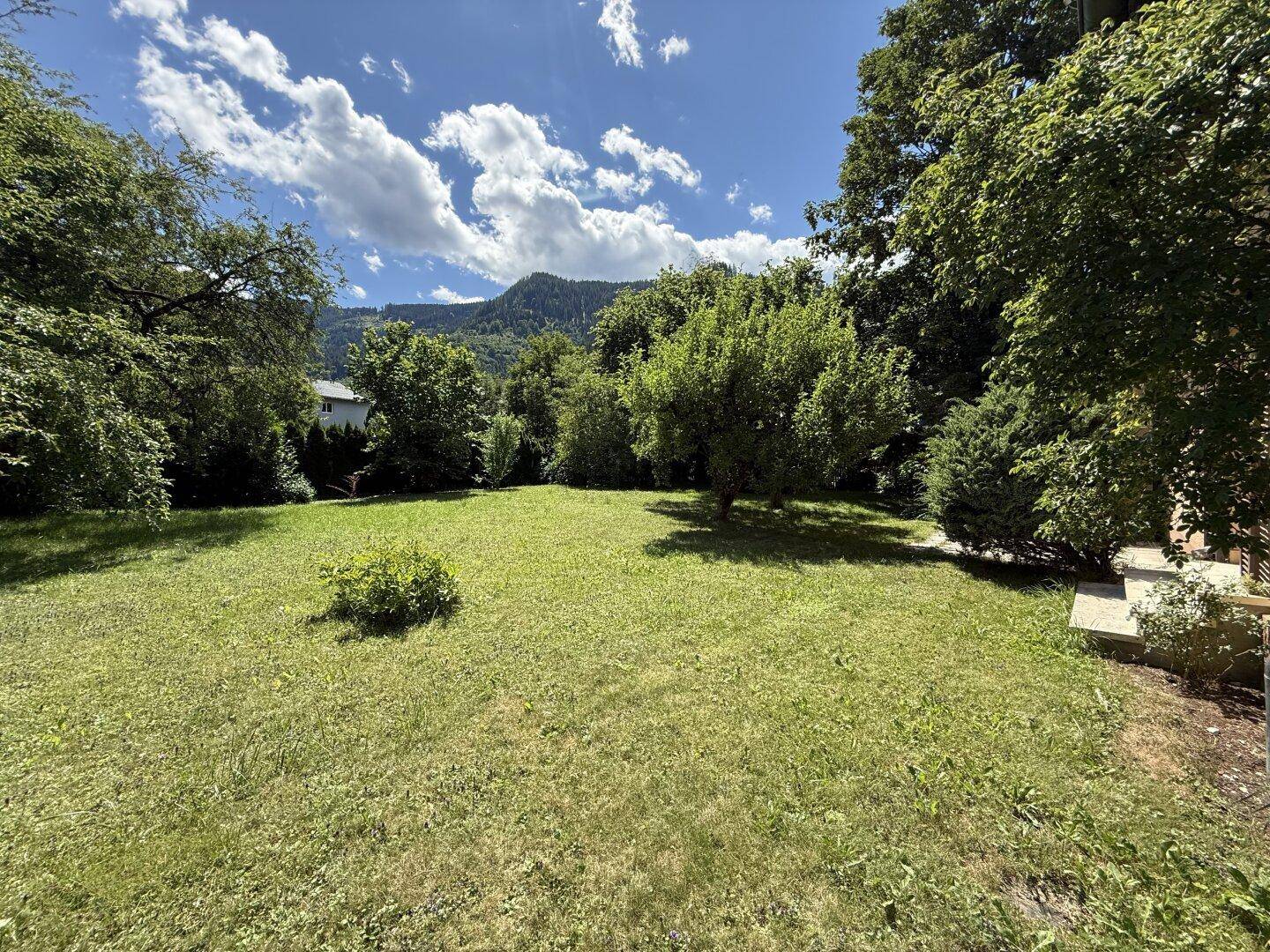 Weitläufiger Garten mit viel Grünfläche, umgeben von Bäumen und einem beeindruckenden Bergpanorama.