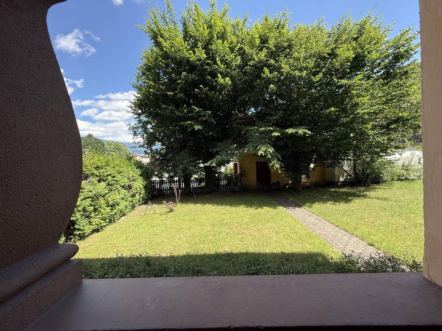 Blick vom überdachten Eingangsbereich in den weitläufigen Garten mit großen Bäumen und Rasenfläche.