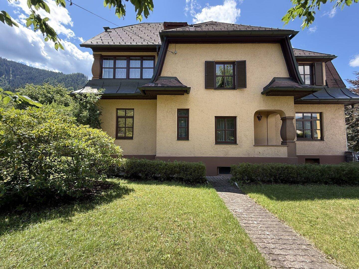Traditionelles Haus mit gelber Fassade, braunen Fensterläden und gepflegtem Vorgarten vor Bergkulisse.
