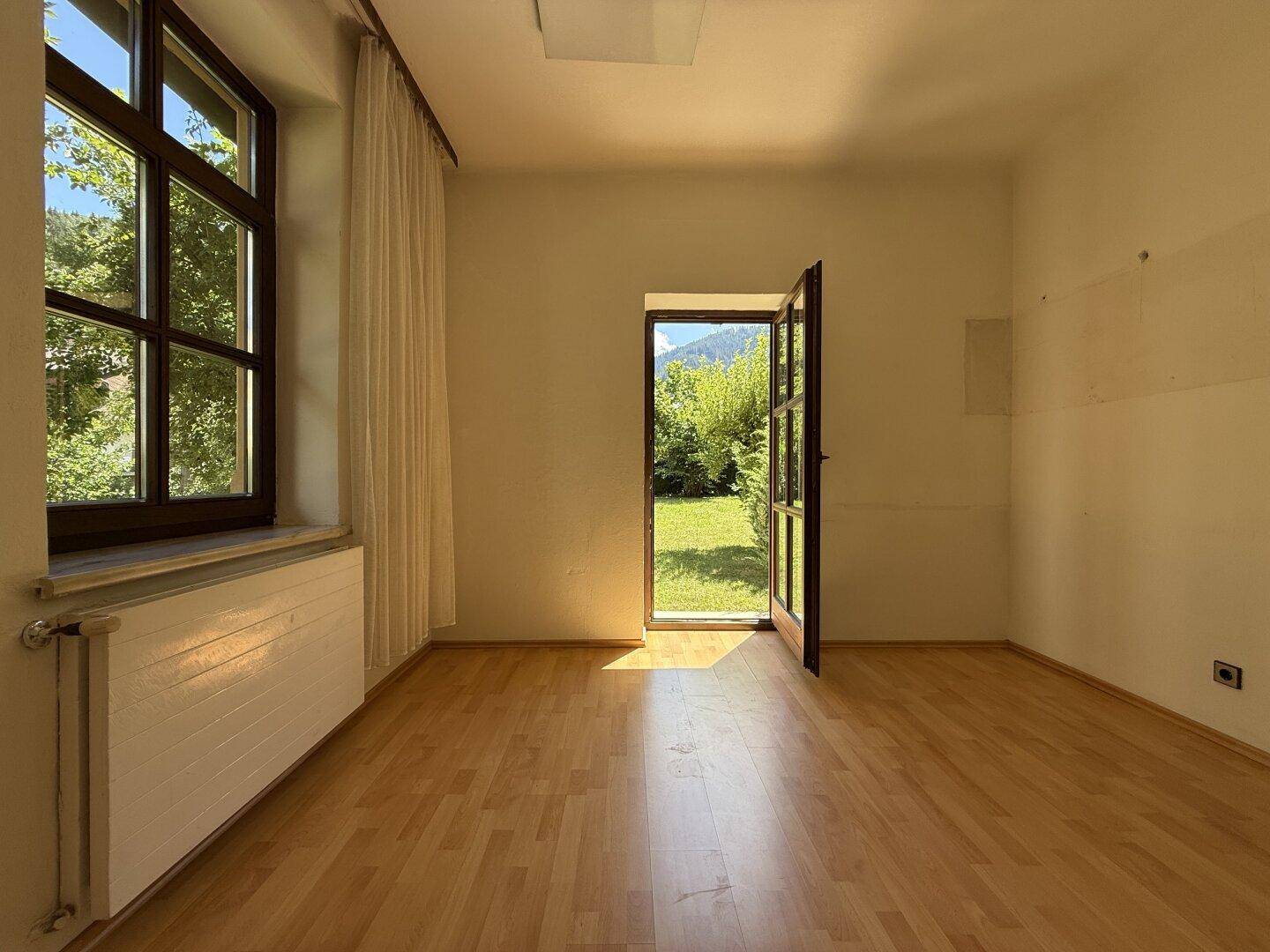 Helles Zimmer mit Holzboden, großem Fenster und direktem Zugang zum grünen Garten.