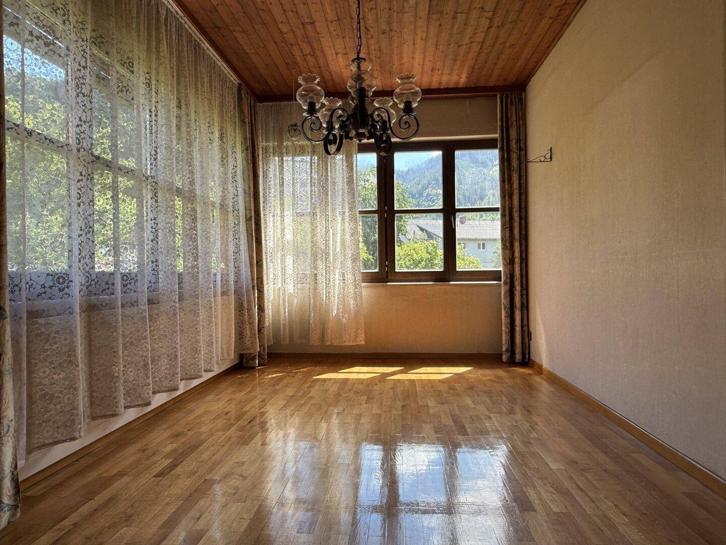 Geräumiges Zimmer mit Holzboden, Holzdecke, großen Fenstern und einem klassischen Kronleuchter.