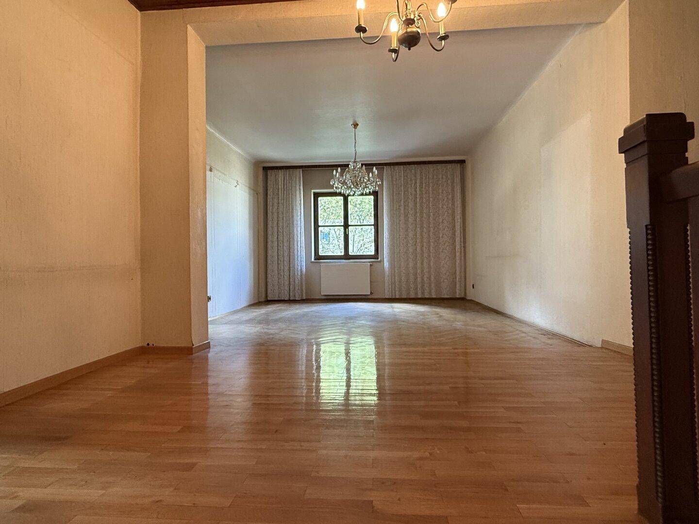 Helles Zimmer mit glänzendem Holzboden, großem Fenster und Kronleuchter, mit Blick auf den Treppenaufgang.