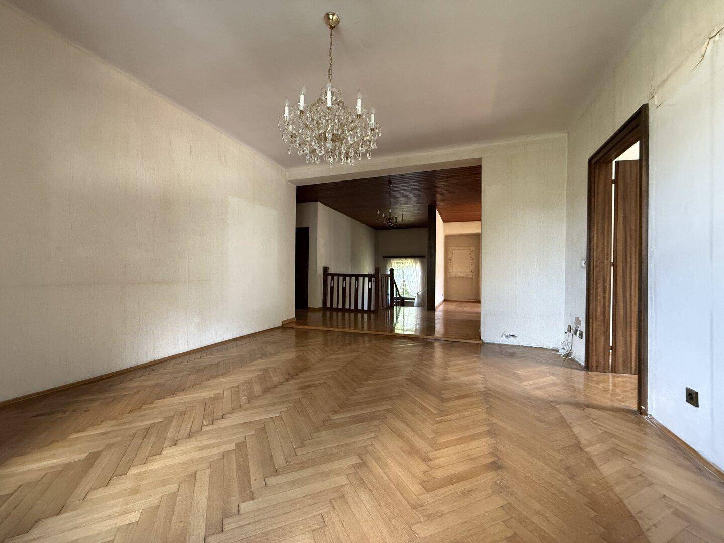 Geräumiges Wohnzimmer mit elegantem Fischgrätparkett und offenem Übergang zum Treppenbereich.