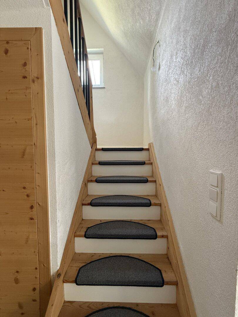 Holztreppe mit Teppichstufen und hellem Geländer, die in das obere Stockwerk führt.