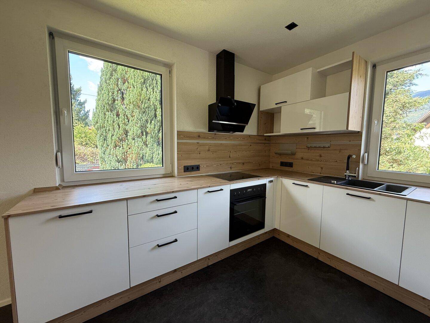 Moderne Küche mit weißen Schränken, Holzdetails und Blick ins Grüne durch große Fenster.