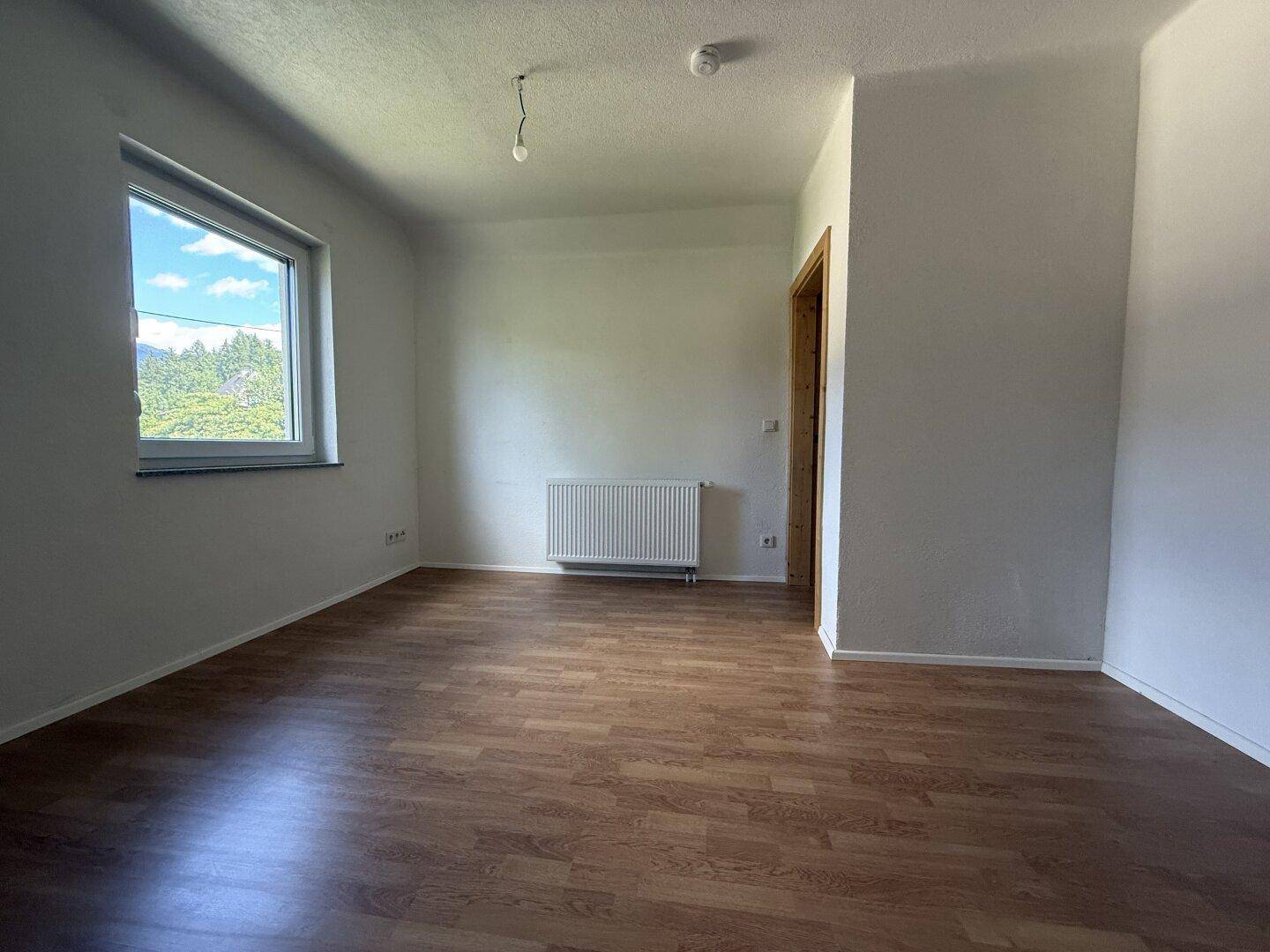 Helles Schlafzimmer mit Laminatboden, Heizkörper und Fenster mit Blick ins Grüne.