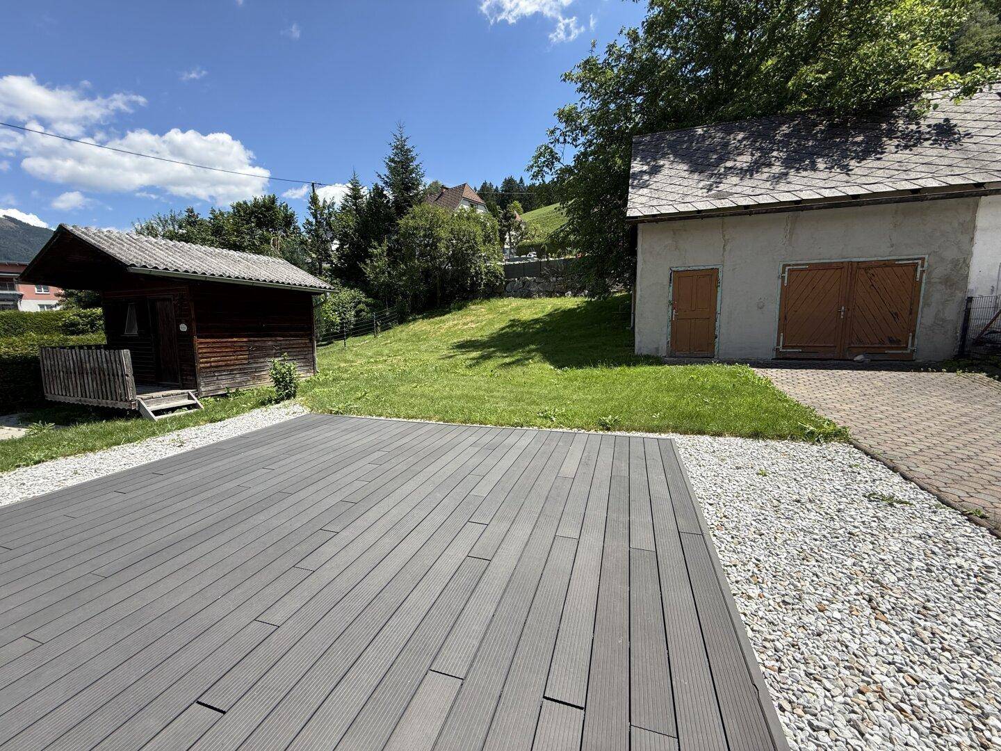 Große Holzterrasse im Gartenbereich mit Blick auf eine alte Scheune und grüne Hügel.