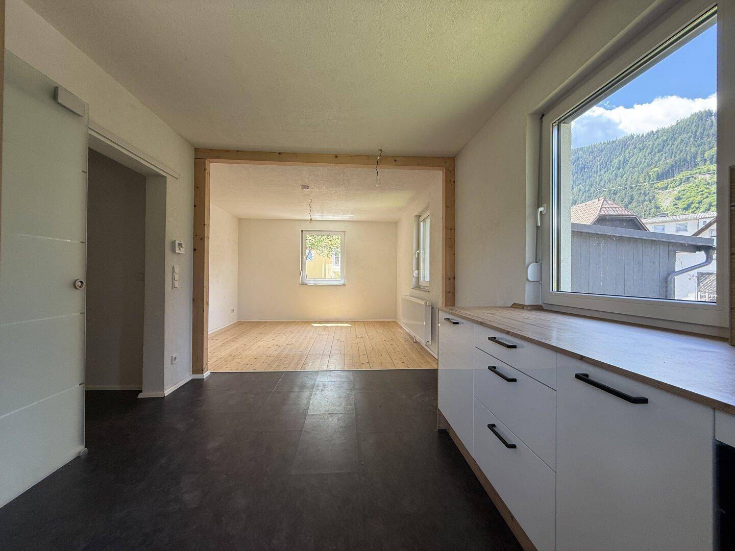 Offener Küchenbereich mit dunklem Boden, der in ein helles Wohnzimmer mit Holzboden übergeht.
