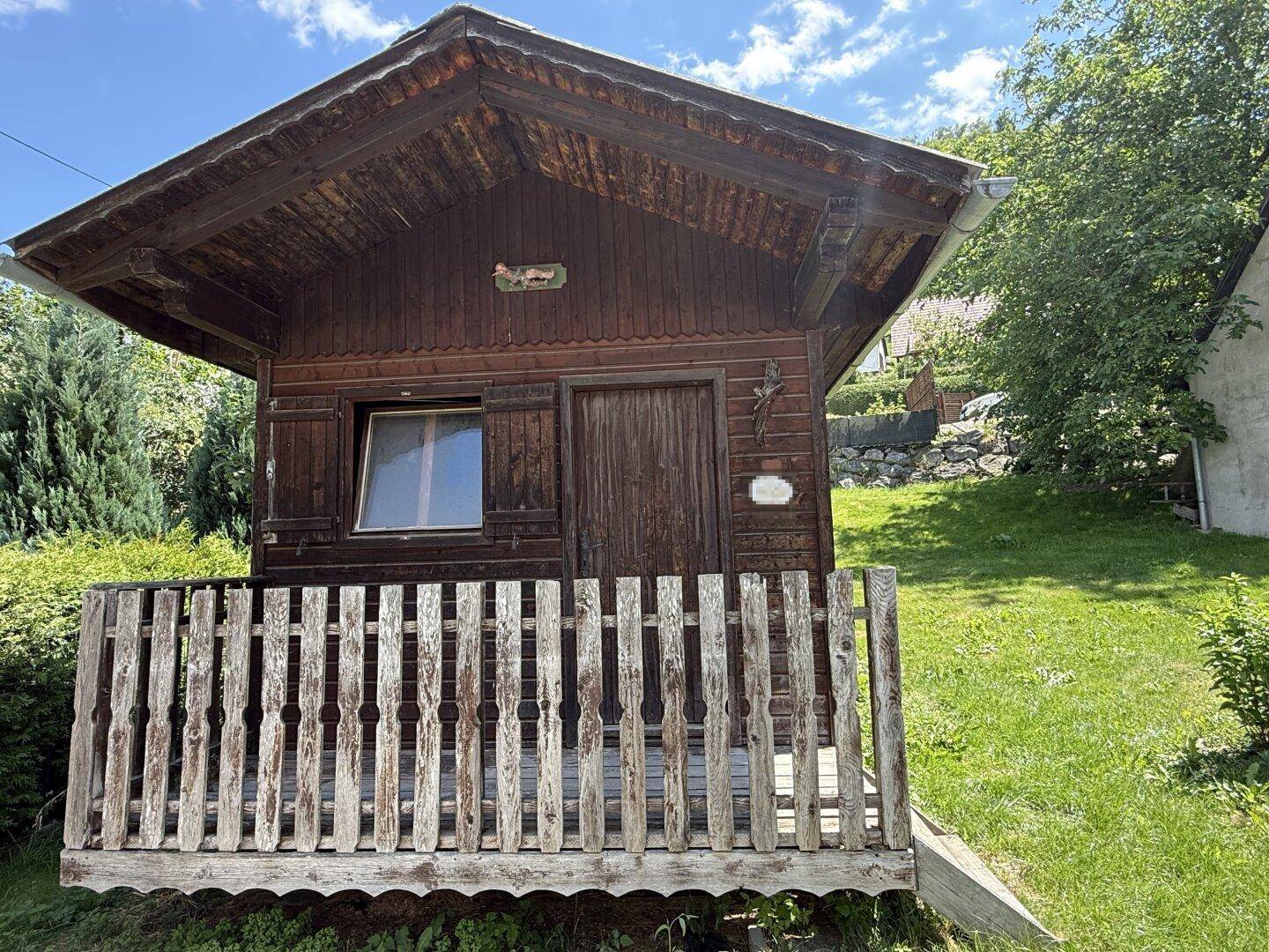 Rustikales Holzgartenhaus mit kleiner Veranda und Fenster in einem grünen Garten.
