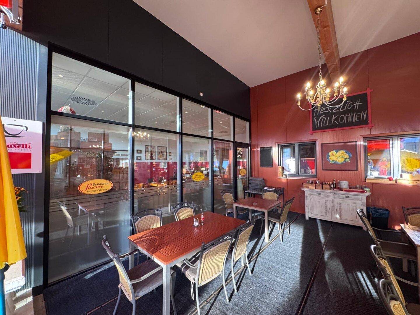 Langer Restaurantbereich mit dunklem Teppichboden, großen Fenstern und einer roten Akzentwand mit Kronleuchter.