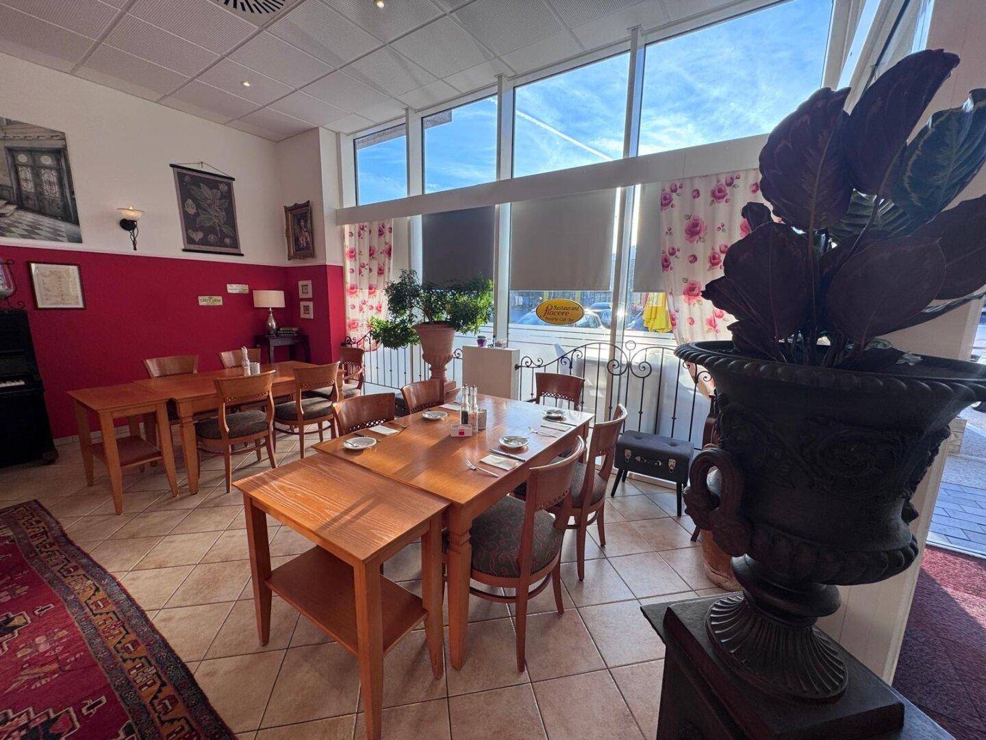 Sonniger Restaurantbereich mit mehreren Holztischen, Fliesenboden und großen Fenstern mit Blick nach draußen.