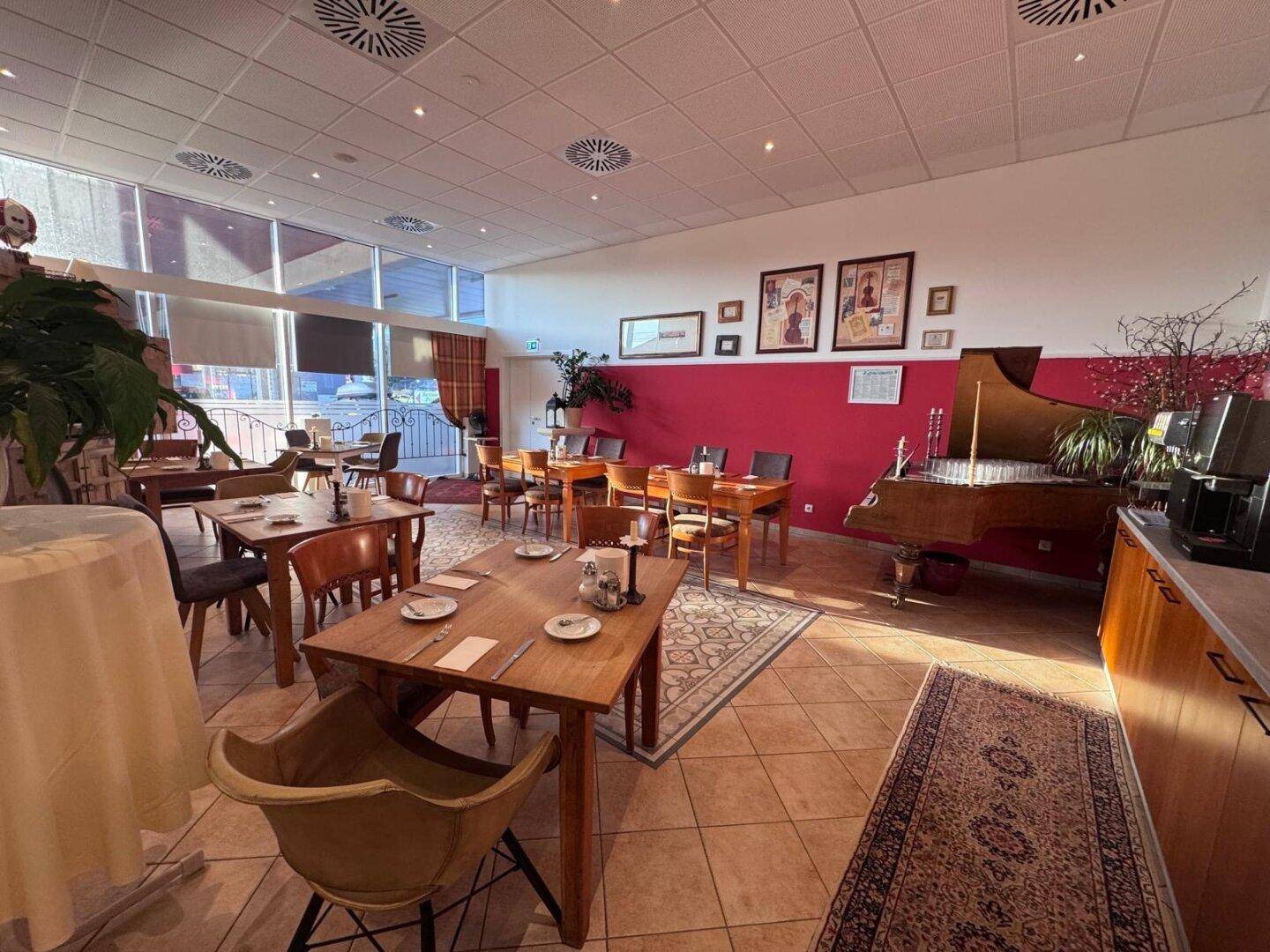 Geräumiger Restaurantbereich mit Holztischen, Fliesenboden und einem eleganten Flügel an der roten Akzentwand.