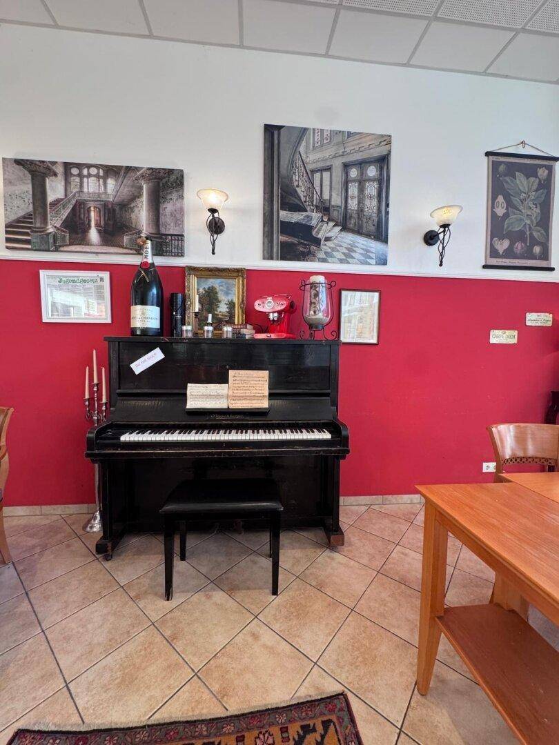 Schwarzes Klavier an einer roten Wand in einem Restaurantbereich, umgeben von Kunstwerken und Wandleuchten.