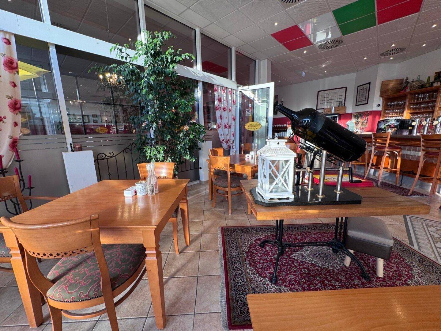 Geräumiger Restaurantbereich mit Holztischen, Fliesenboden und einer großen Weinflasche als Dekoration.
