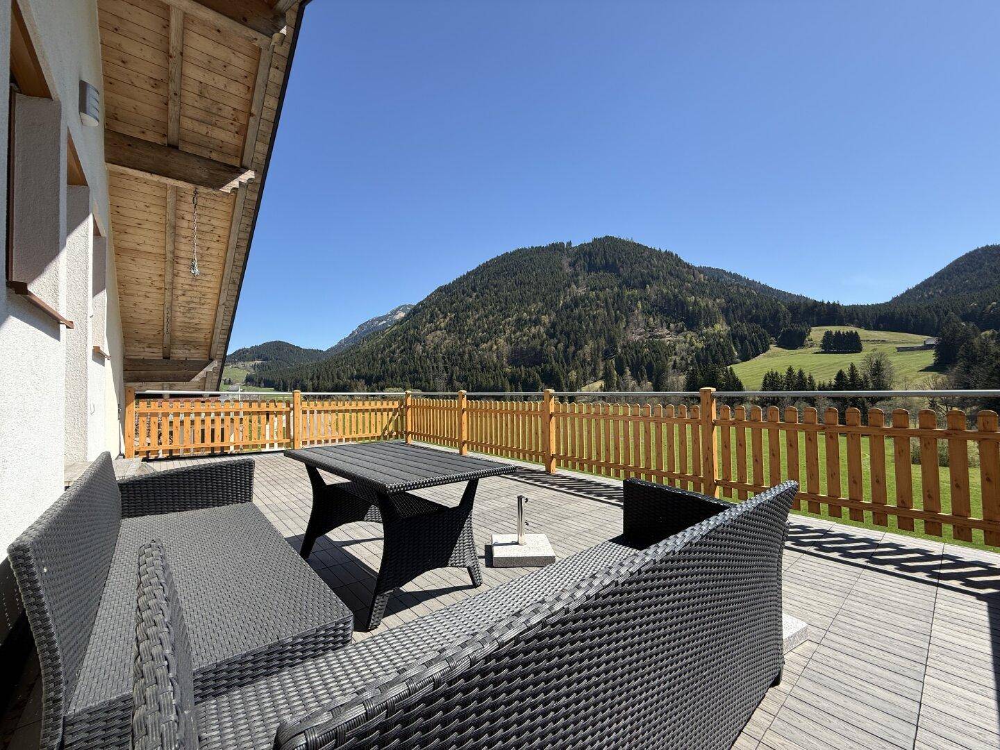 Möblierte Terrasse mit Rattanmöbeln und Panoramablick auf die bewaldeten Berge unter strahlendem Sonnenschein.