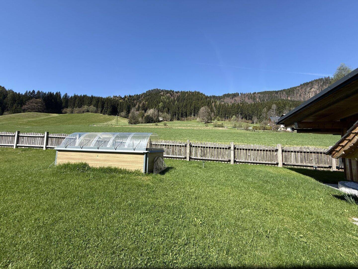 Großer grüner Gartenbereich mit einem kleinen Gewächshaus und Blick auf die Natur und Wälder.