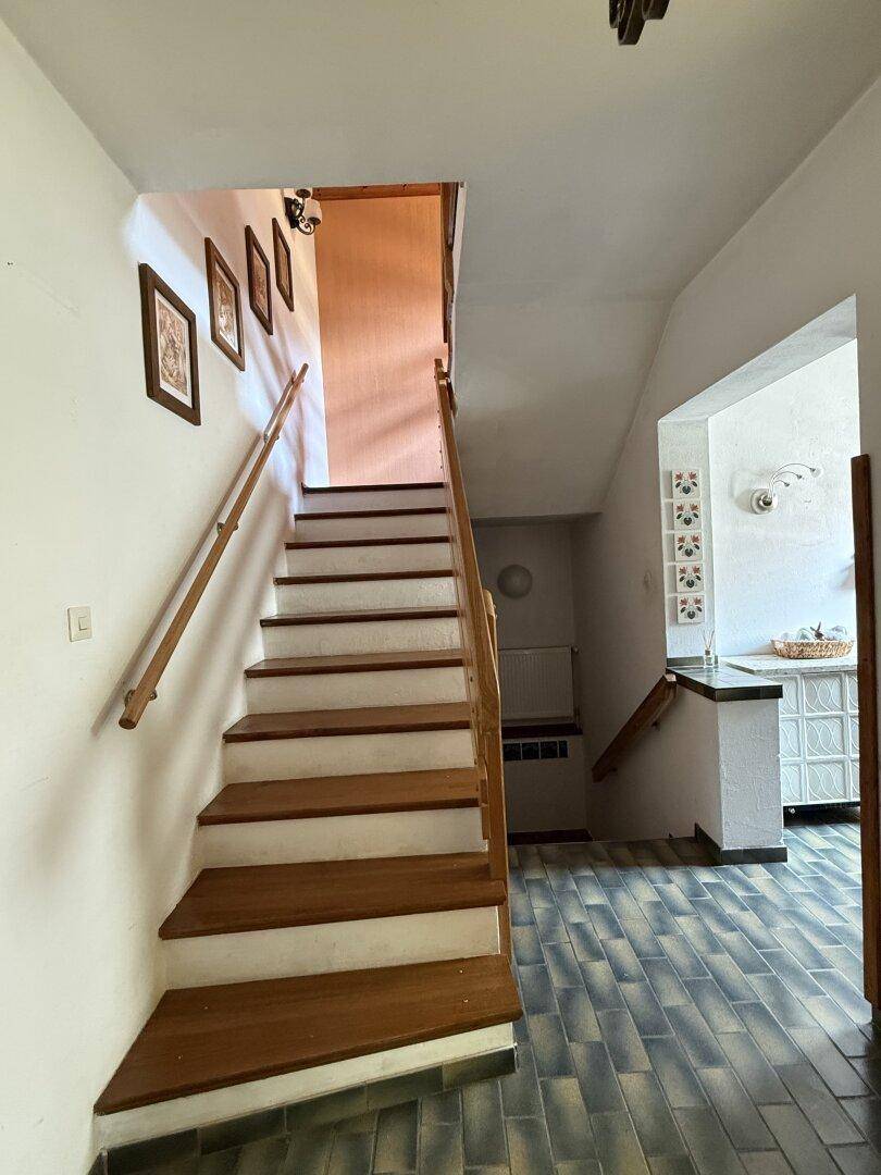 Holztreppe mit Geländer führt in die obere Etage, im Vordergrund gefliester Boden.