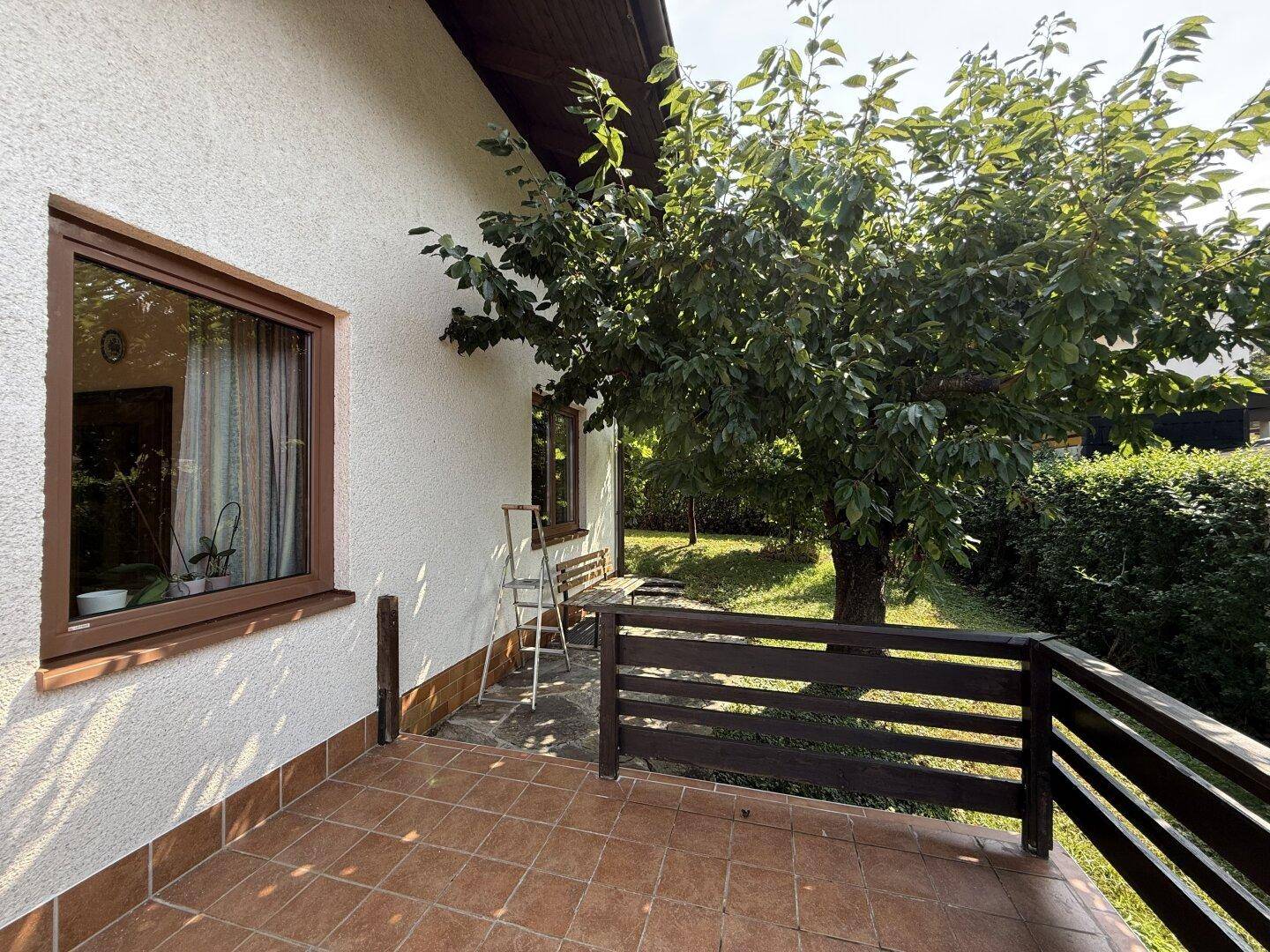 Seitlicher Blick auf die geflieste Terrasse mit schattenspendendem Baum und Hausfassade.