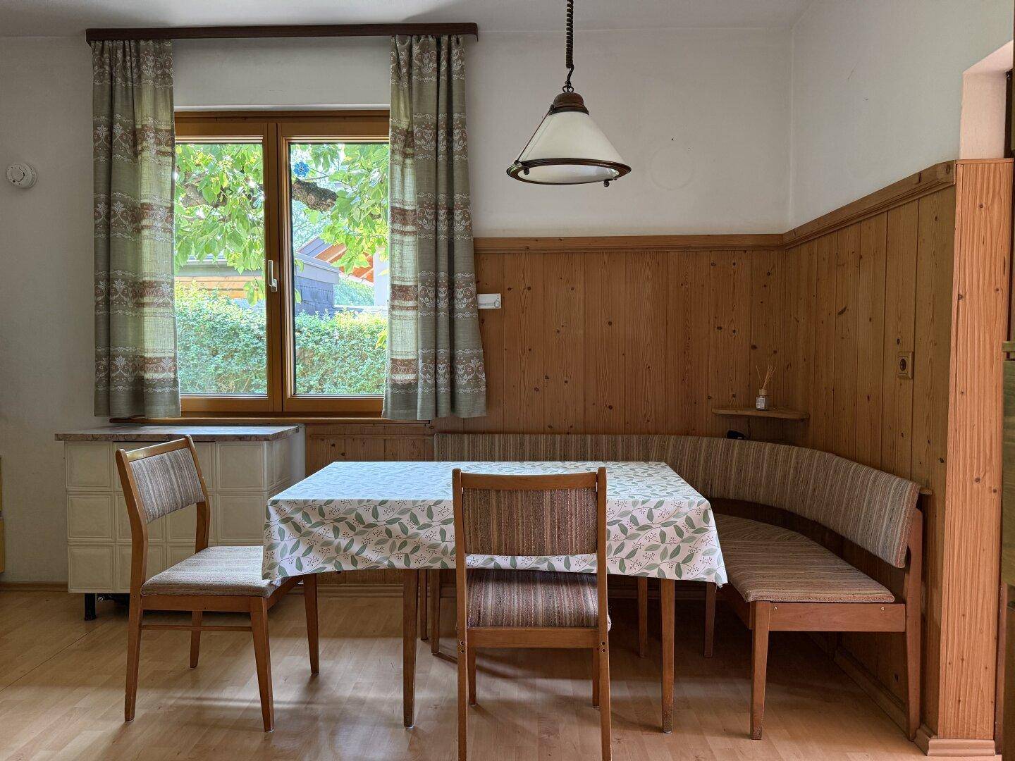 Gemütlicher Essbereich mit traditioneller Eckbank, Holztisch und Blick ins Grüne durch das Fenster.