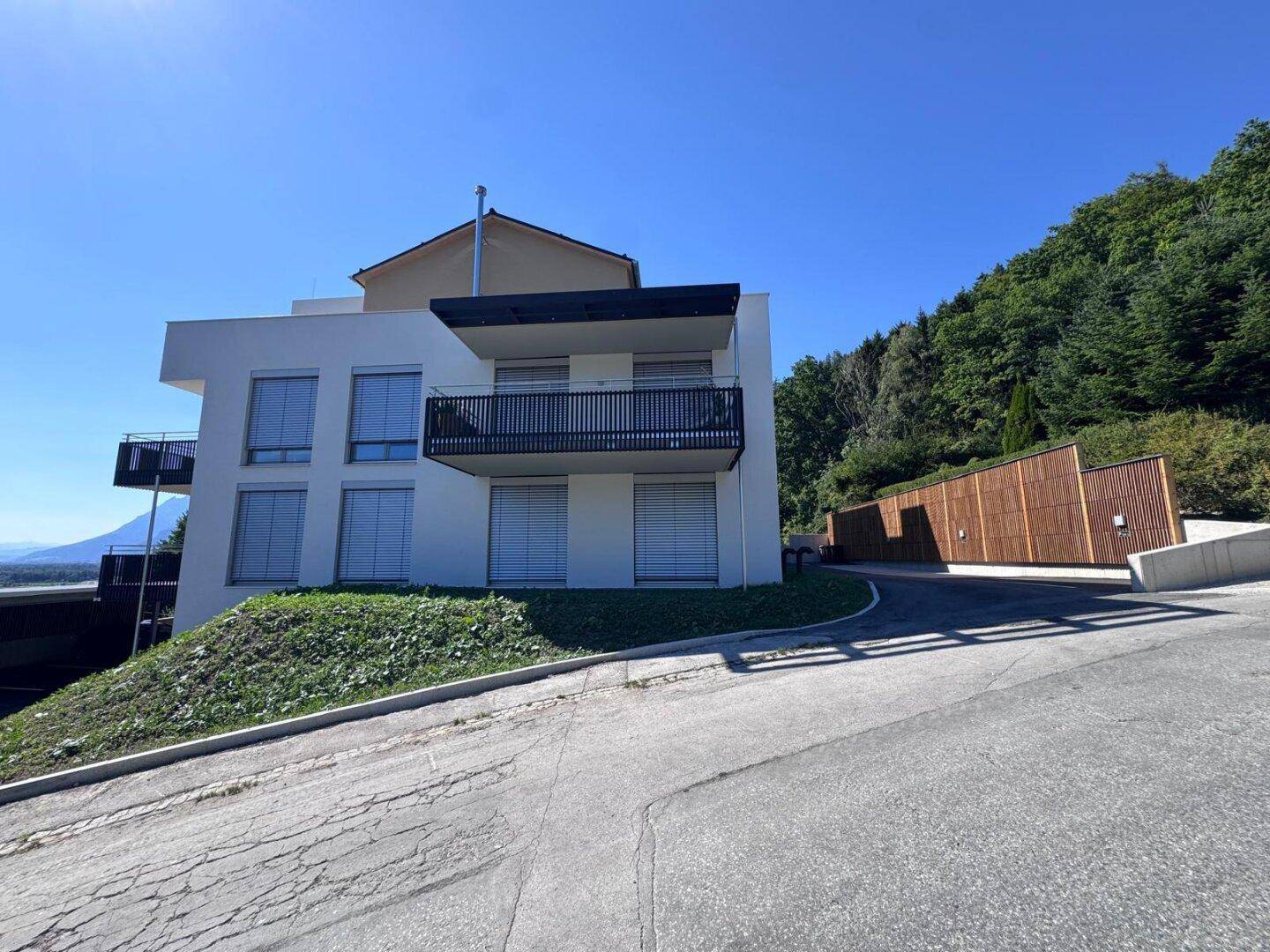 Modernes Wohngebäude mit Balkon und Carport, umgeben von grüner Natur und Bergen.