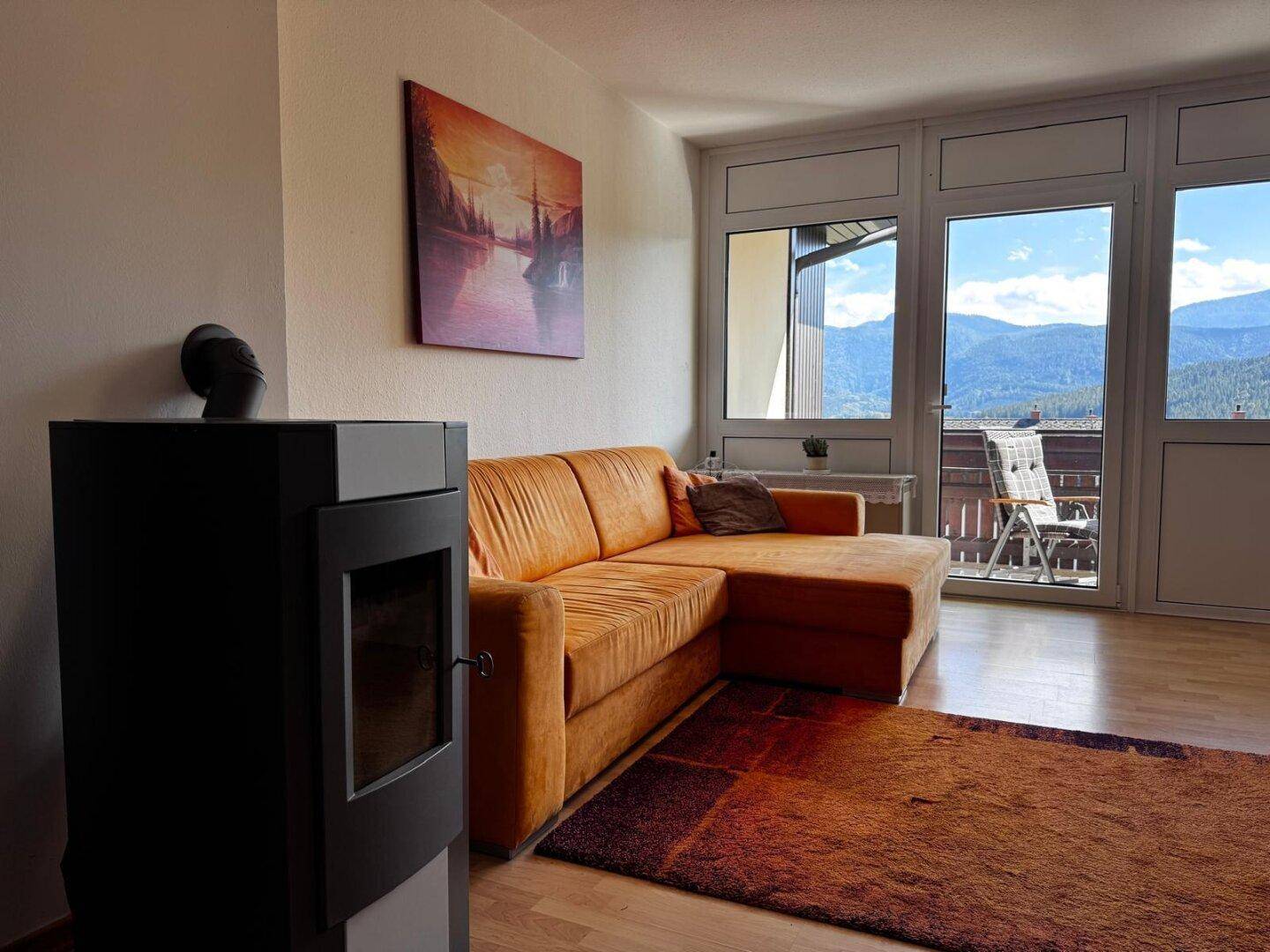Helles Wohnzimmer mit gemütlichem Sofa, modernem Holzofen und Zugang zum Balkon mit Bergblick.