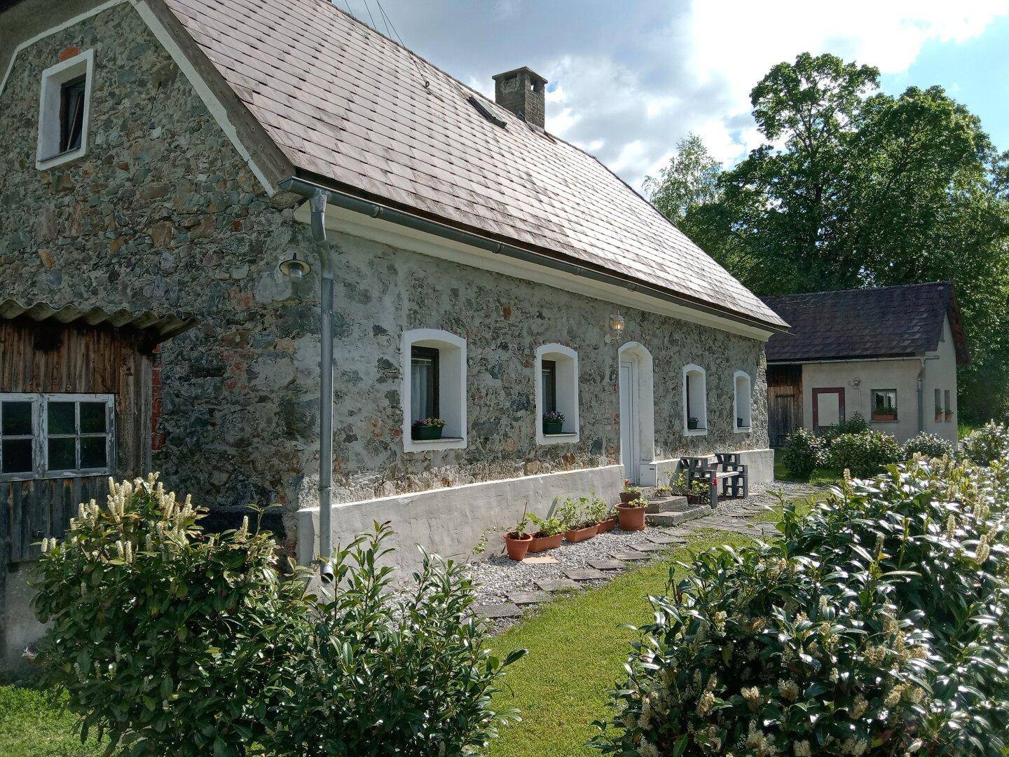 Seitenansicht des Steinhauses mit kleinem Gartenbereich und Topfpflanzen, umgeben von grüner Natur.