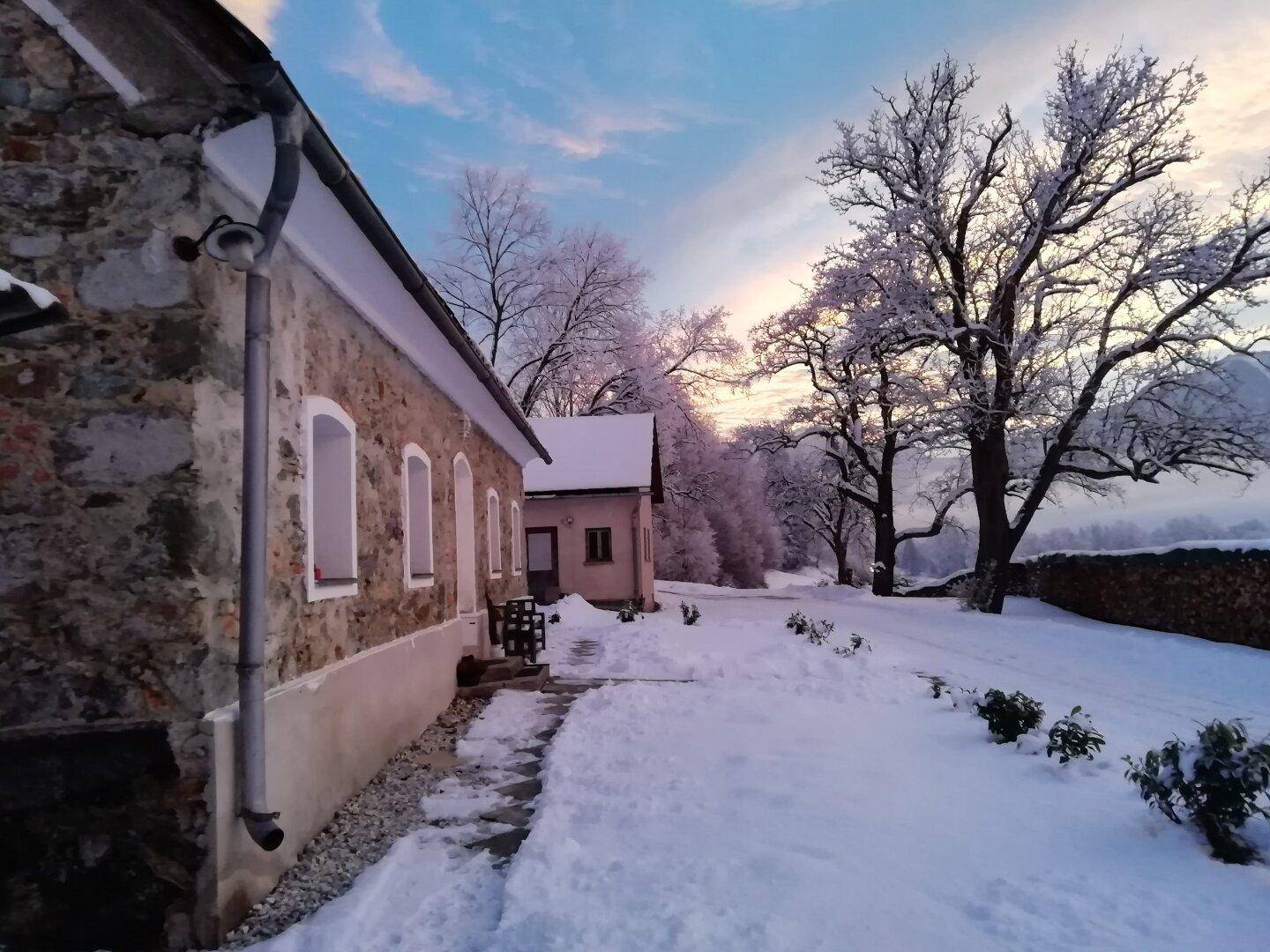 Winterliche Ansicht des Steinhauses mit verschneiter Landschaft und frostbedeckten Bäumen bei Sonnenuntergang.