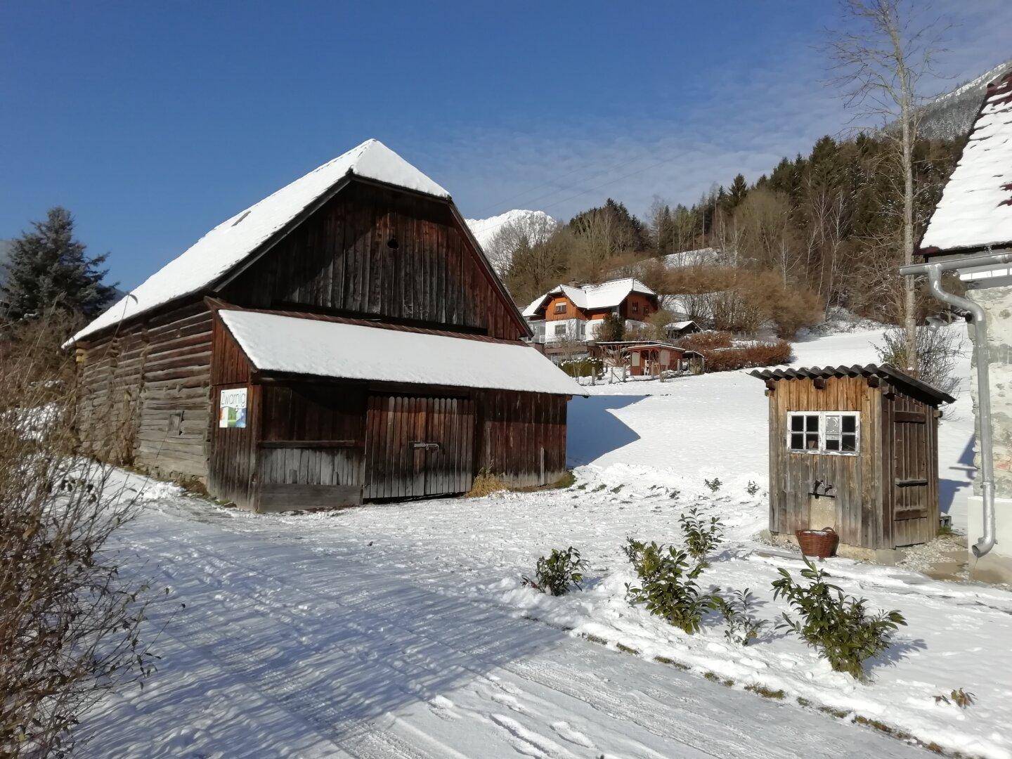 Verschneite Scheune und kleiner Holzschuppen in einer idyllischen Winterlandschaft mit Bergen im Hintergrund.