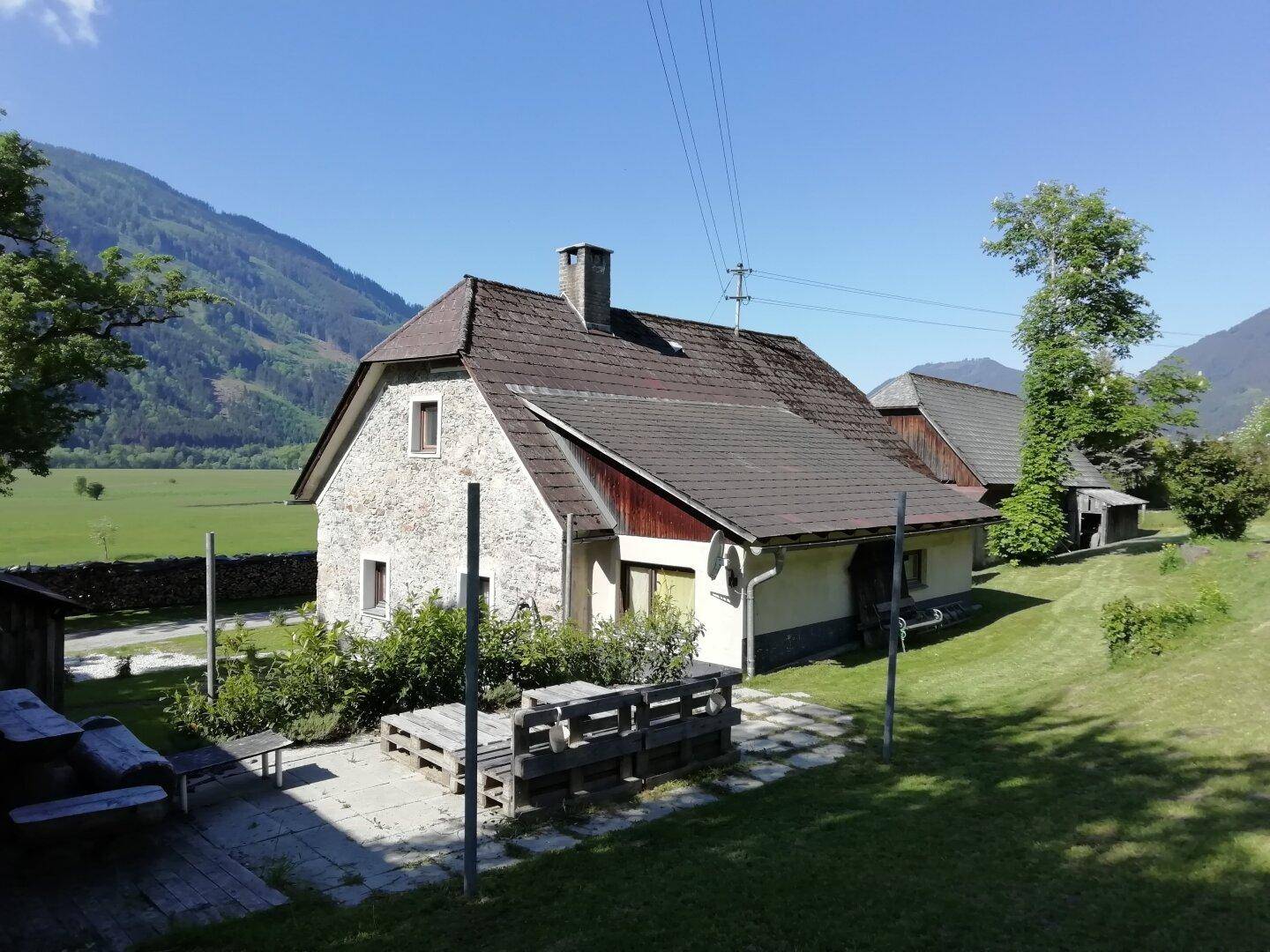 Steinhaus mit einer gepflasterten Terrasse und Sitzgelegenheiten, umgeben von weiten Wiesen und Bergen.