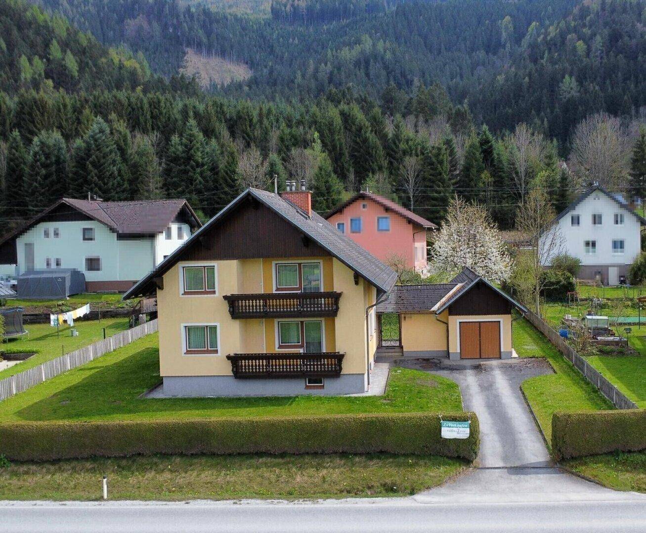 Gelbes Einfamilienhaus mit zwei Balkonen, umgeben von einem gepflegten Garten und Wald im Hintergrund.