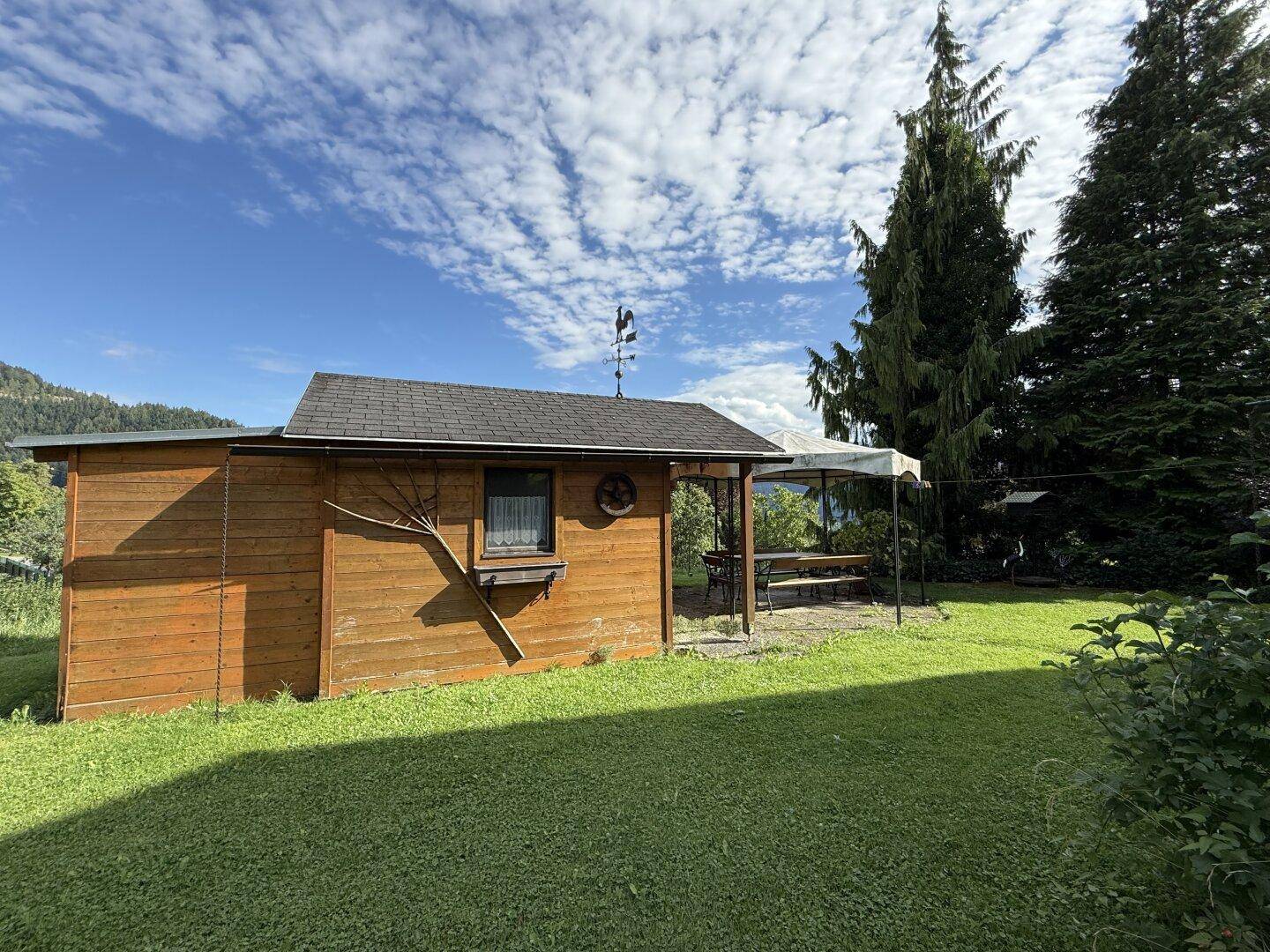 Kleines Gartenhaus aus Holz mit überdachtem Sitzbereich im weitläufigen Garten unter blauem Himmel.