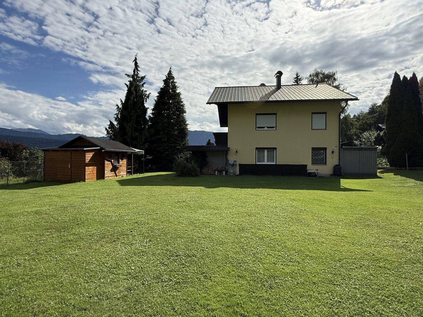 Rückansicht des Hauses mit weitläufigem Rasen, Gartenhaus und Bergblick in der Ferne.