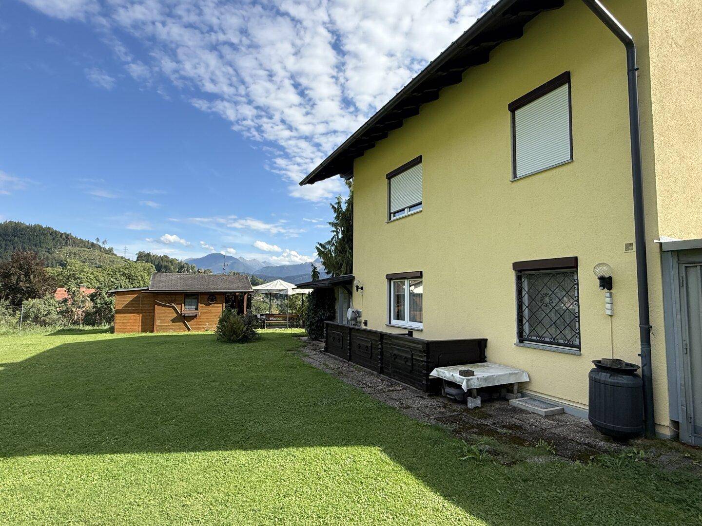 Seitenansicht des Hauses mit großem Garten, Holzschuppen und Blick auf die Berge im Hintergrund.