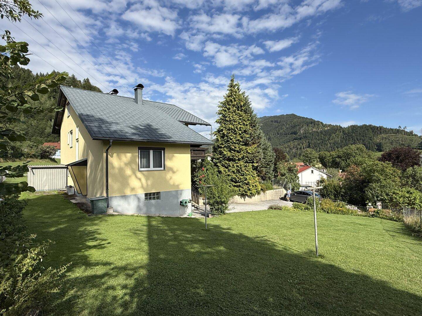Seitenansicht des Hauses mit gepflegtem Rasen und Blick auf die umliegende Hügellandschaft unter blauem Himmel.