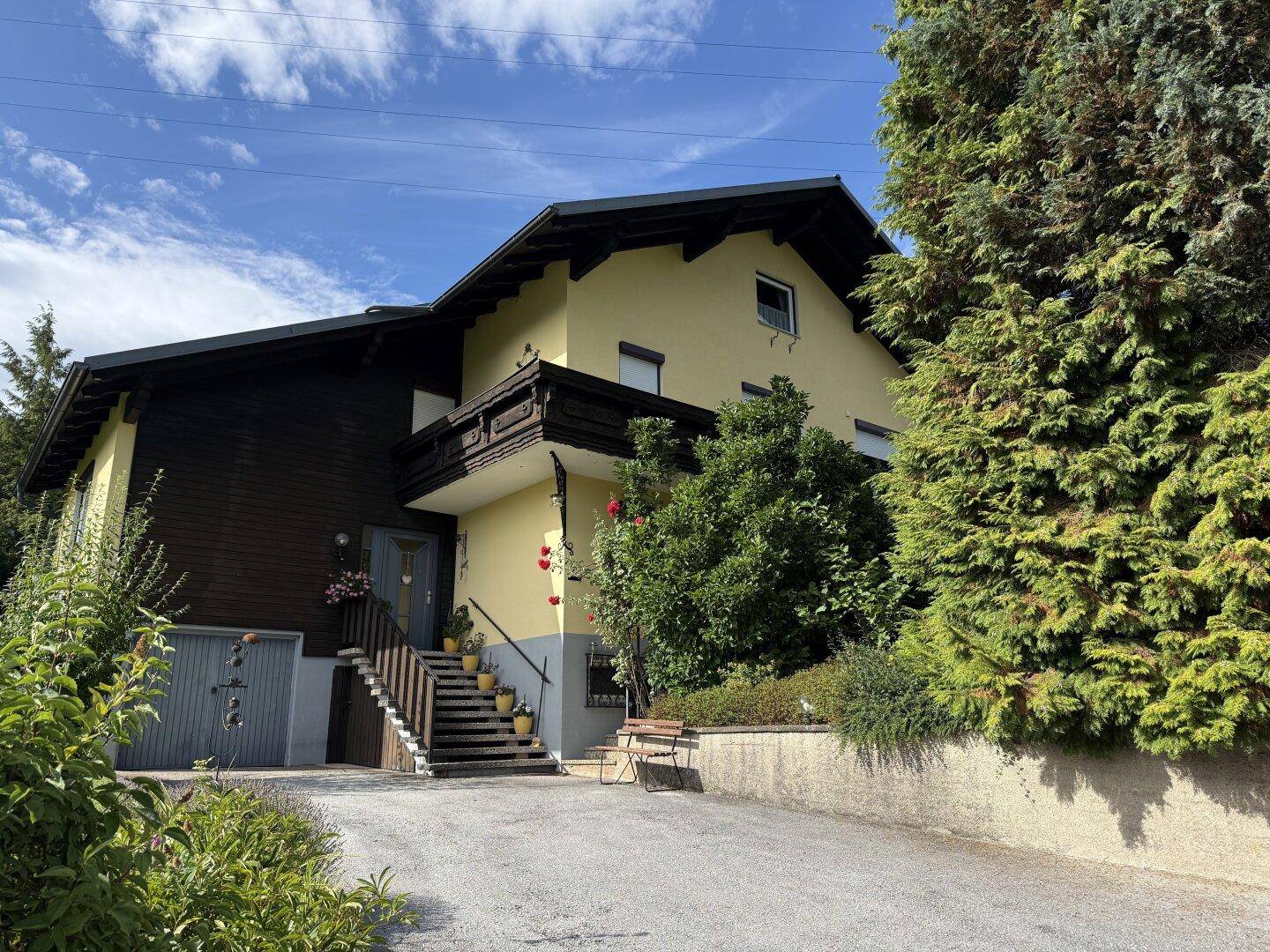 Eingangsbereich des Hauses mit Treppe, Balkon und Zufahrt, umgeben von viel Grün und Bäumen.