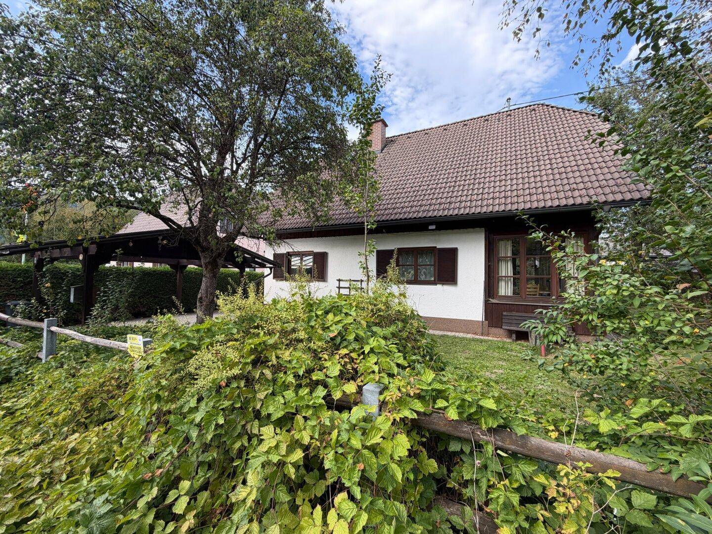 Charmantes Einfamilienhaus mit Satteldach, umgeben von üppigem Grün und einem Carport.