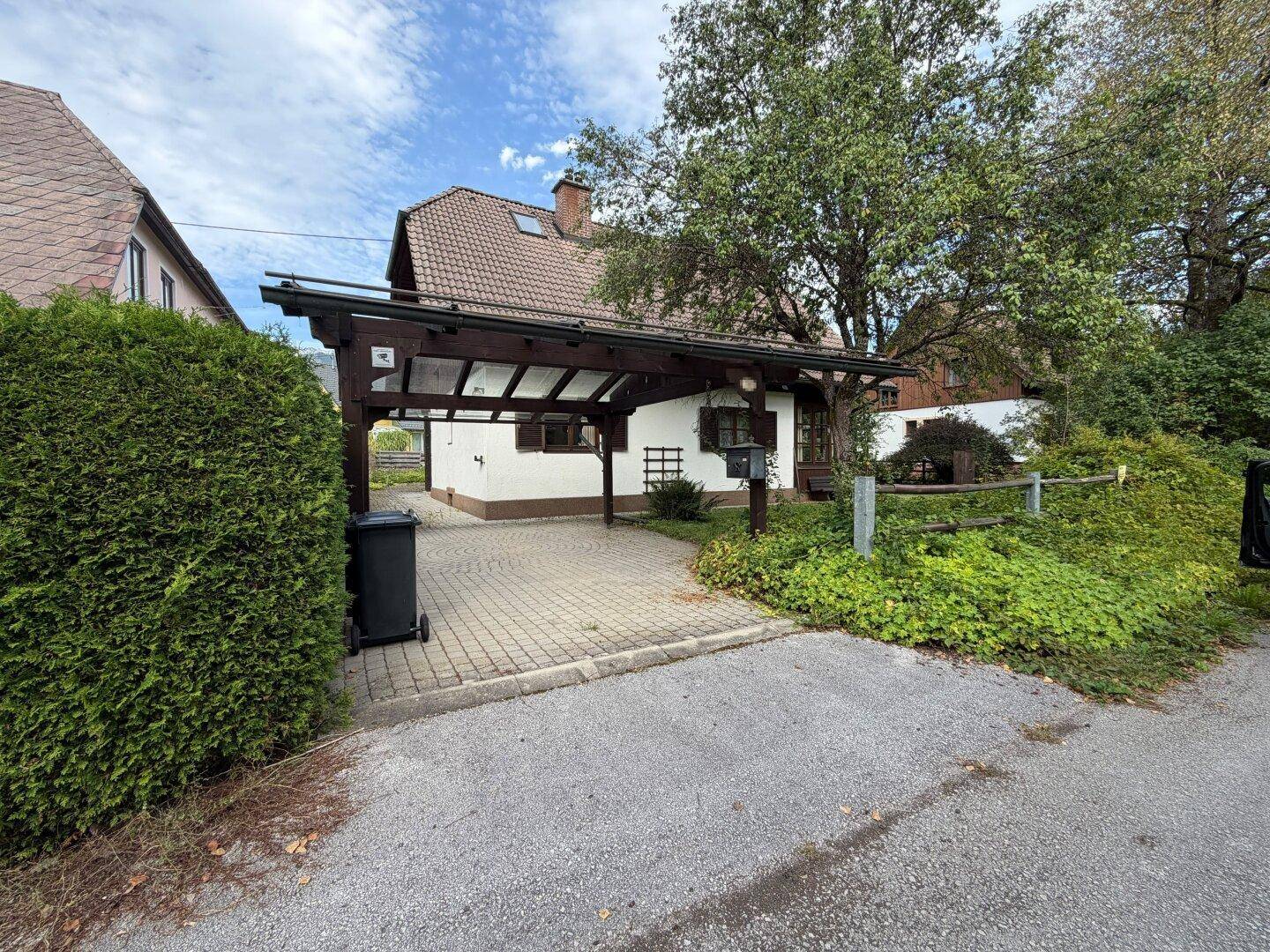 Ansicht des Hauses mit Carport und gepflasterter Einfahrt, umgeben von grüner Bepflanzung.