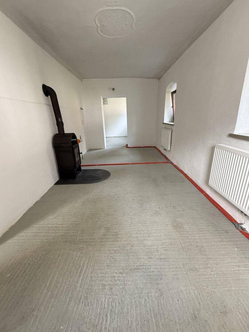Renovierungsbedürftiger Raum mit einem Holzofen und freiliegenden Rohren, bereit für neue Bodenbeläge.