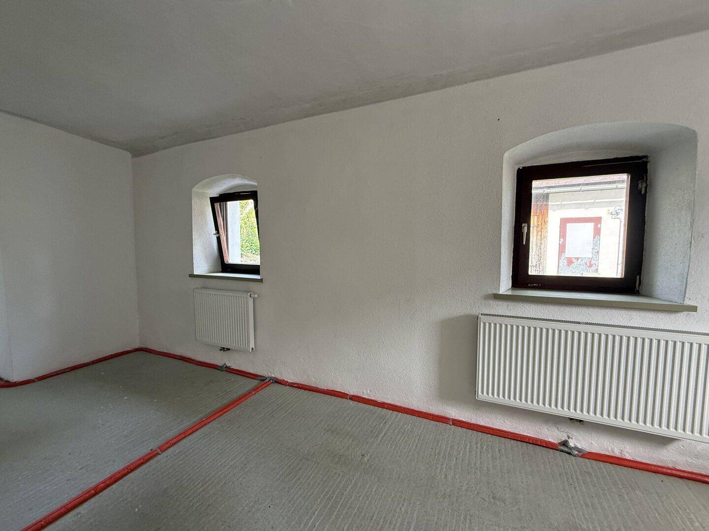 Renovierungsbedürftiger Raum mit zwei Fenstern und Heizkörpern, die auf neue Bodenbeläge warten.
