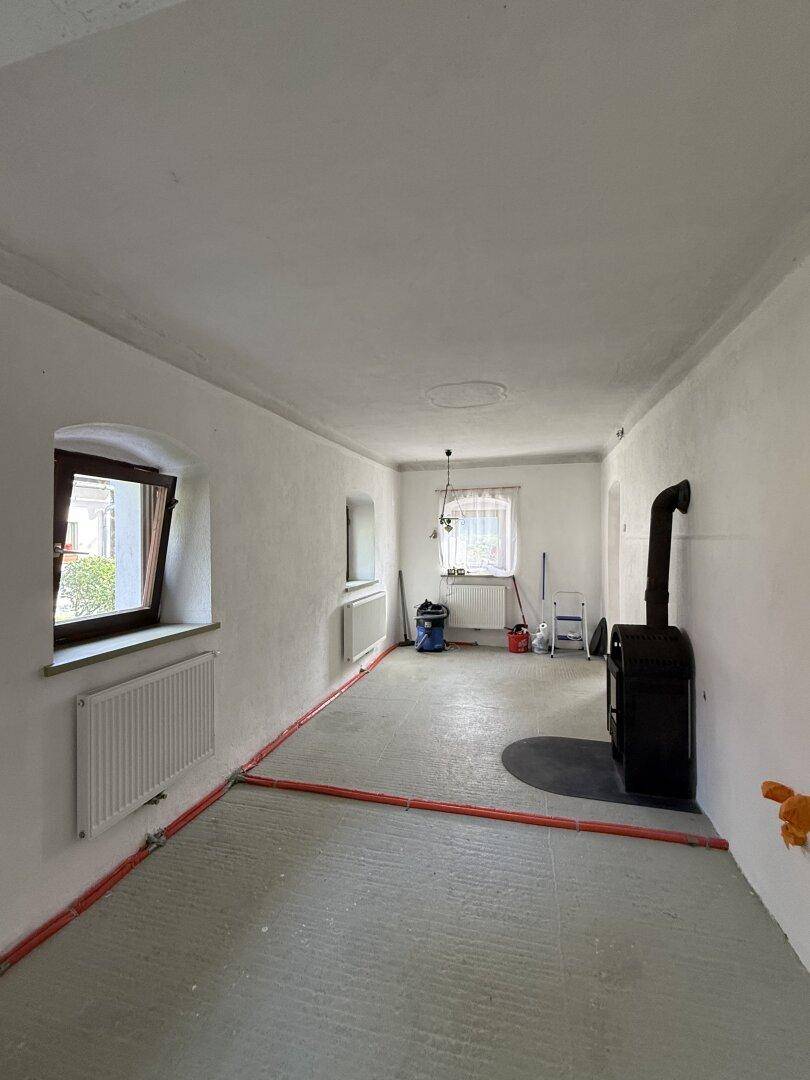 Geräumiger Raum in Renovierung mit Holzofen, Fenstern und sichtbaren Heizungsrohren.
