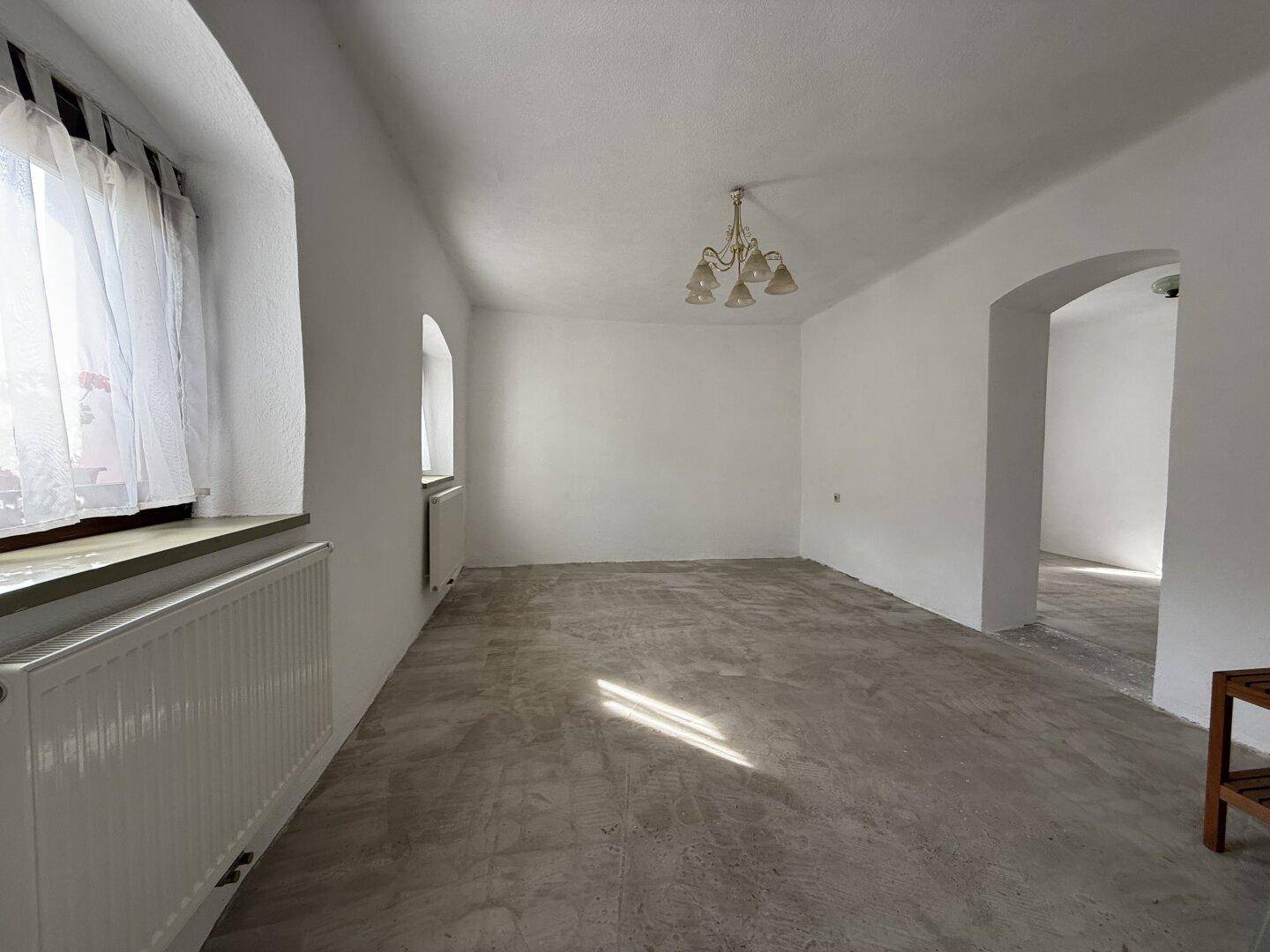 Heller, leerer Raum mit weiß gestrichenen Wänden, Betonboden und einem klassischen Kronleuchter.