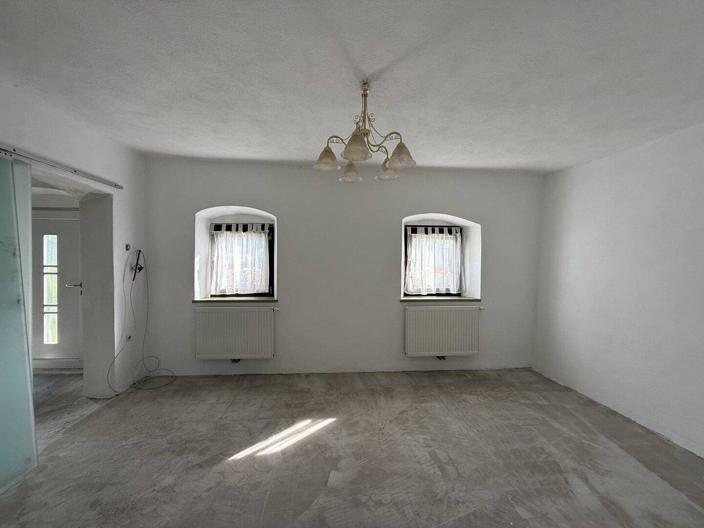 Leerer Raum mit zwei Fenstern, weiß gestrichenen Wänden und einem klassischen Deckenleuchter.