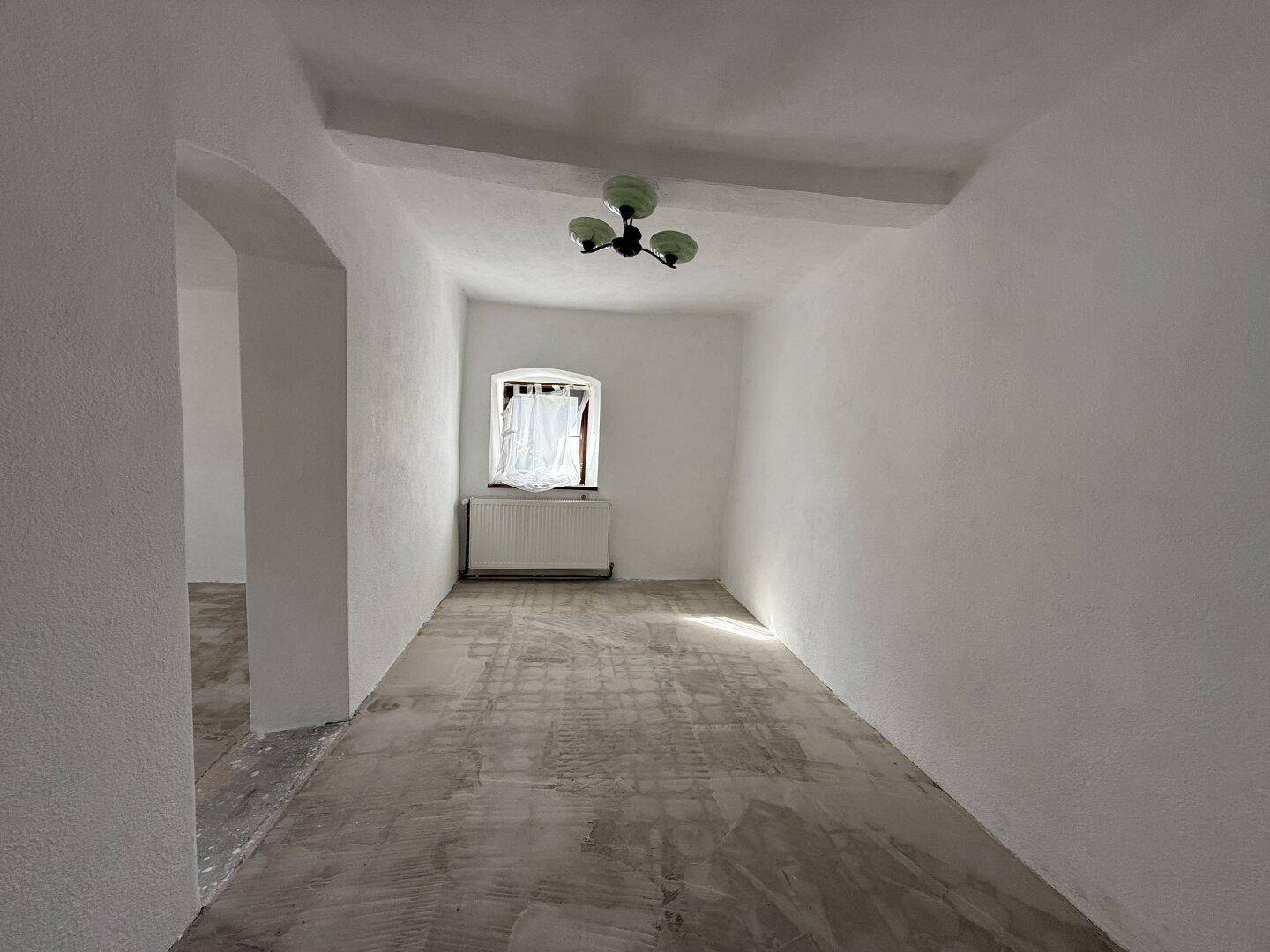 Kleiner, heller Raum mit einem Fenster, weiß gestrichenen Wänden und einem einfachen Deckenlicht.