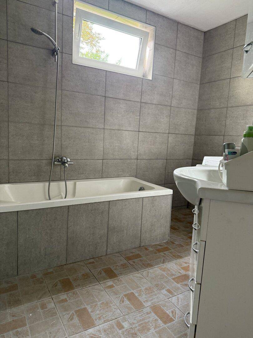 Badezimmer mit grauen Fliesen, Badewanne mit Duschfunktion und einem Waschbeckenunterschrank.