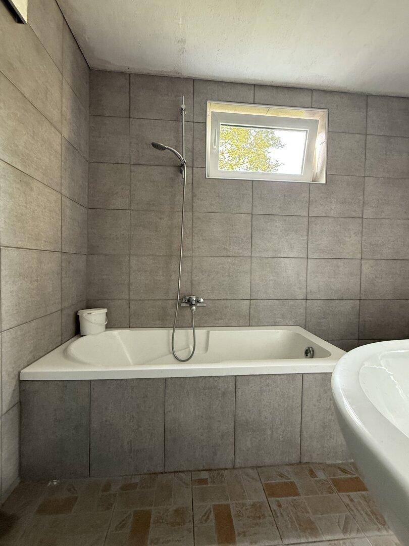 Badezimmer mit grauen Wandfliesen, Badewanne mit Duscharmatur und einem Waschbecken.