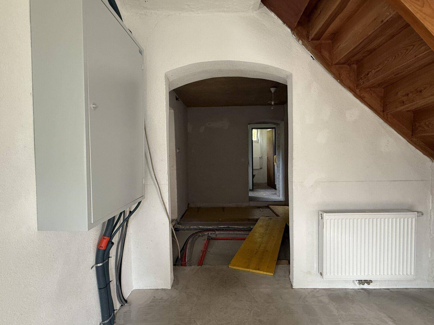 Eingangsbereich in Renovierung mit sichtbaren Leitungen, einem Sicherungskasten und einer Holztreppe.