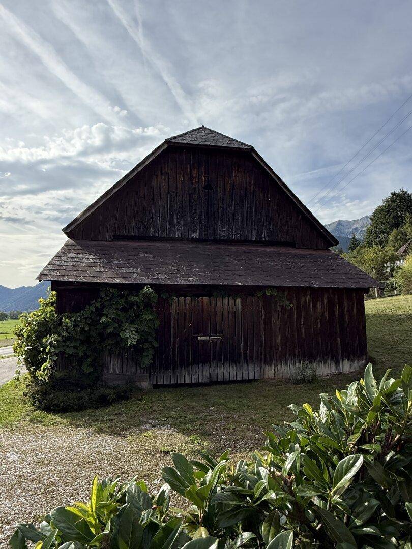 Rustikales Holzgebäude mit dunklem Dach und grüner Vegetation im ländlichen Umfeld mit Bergblick.