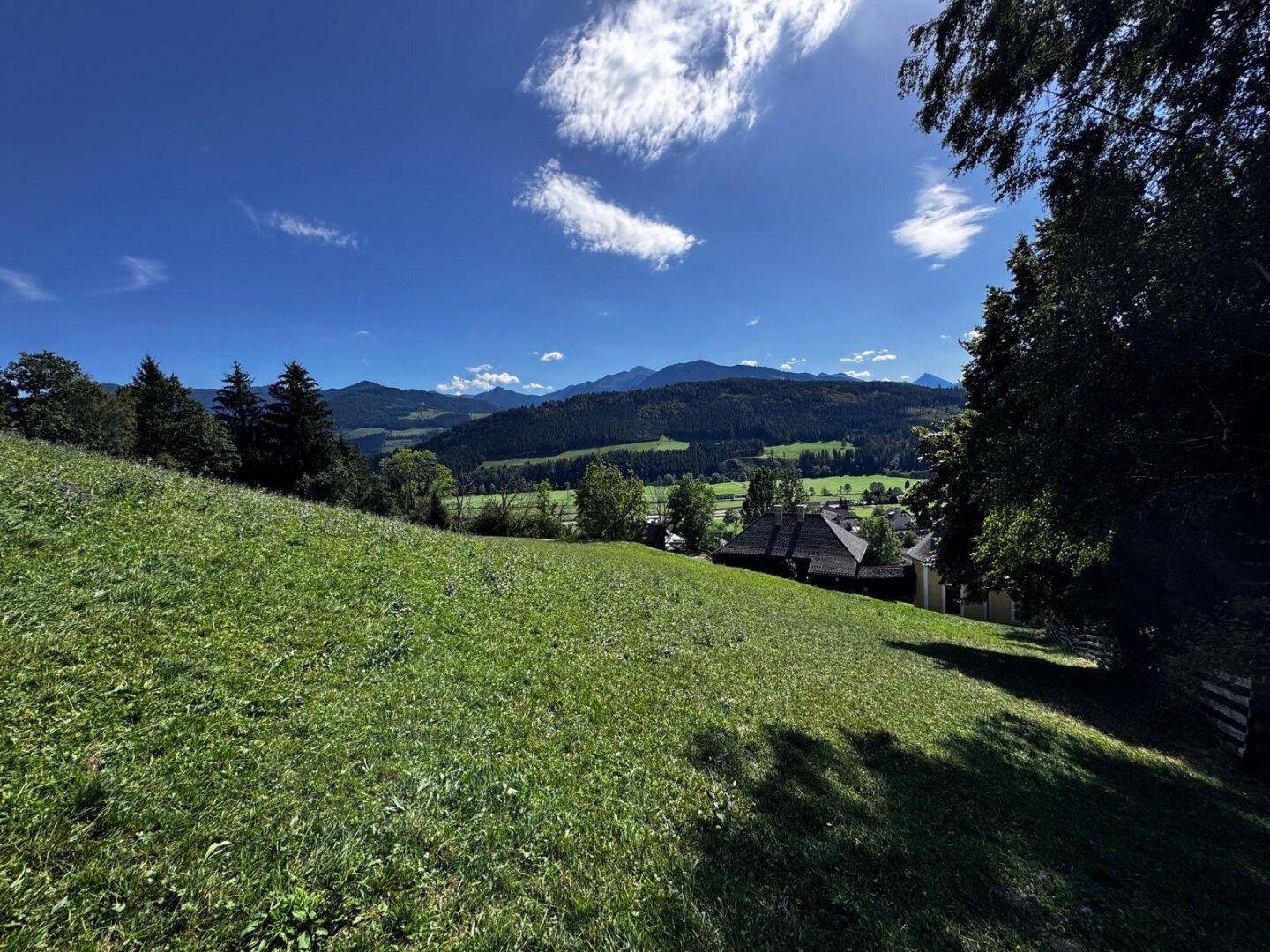 Sonniger Blick über eine grüne Wiese auf ein kleines Dorf und eine weite Berglandschaft am Horizont.
