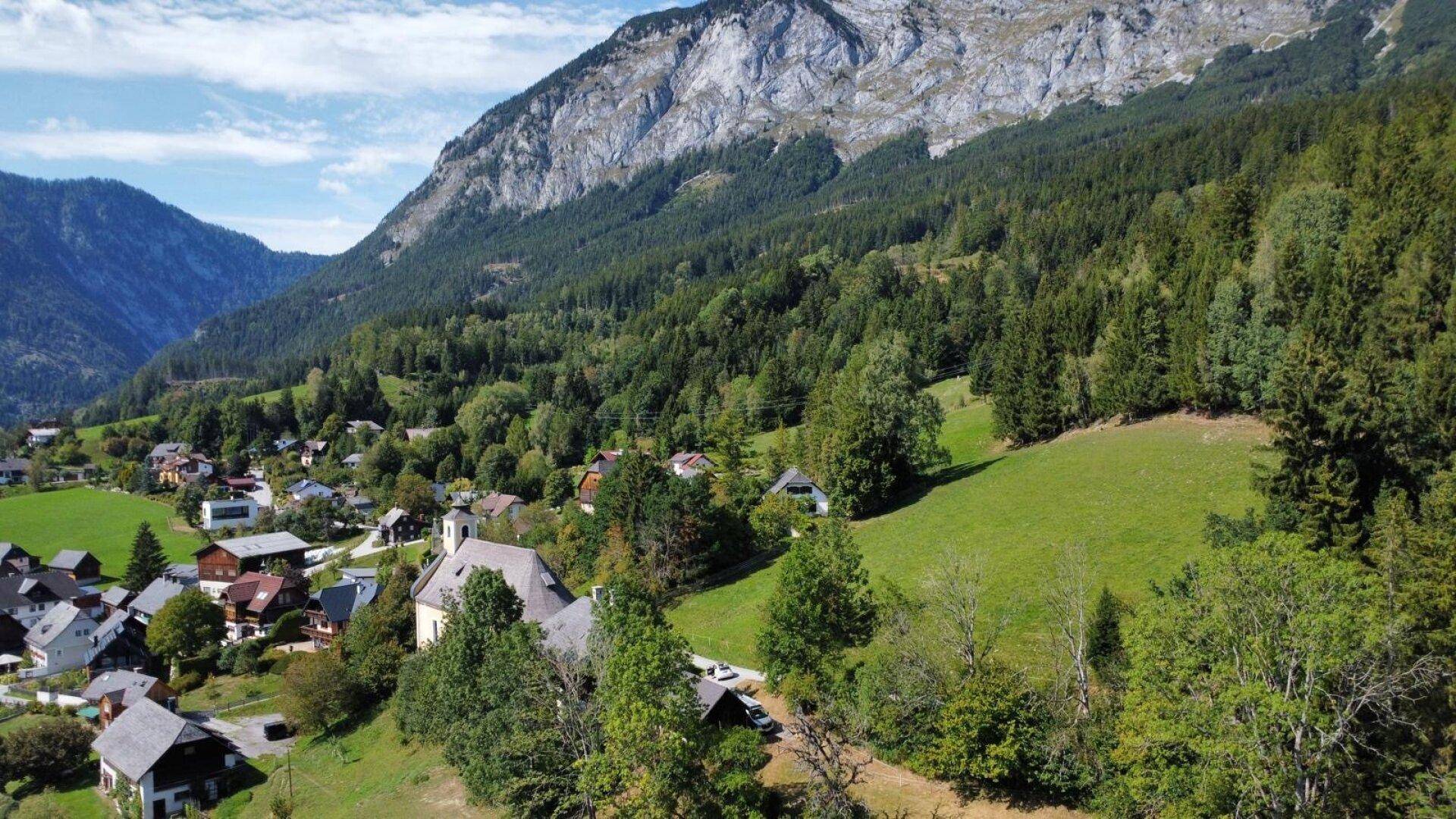 Luftaufnahme eines idyllischen Dorfes eingebettet in ein grünes Tal, umgeben von bewaldeten Bergen.